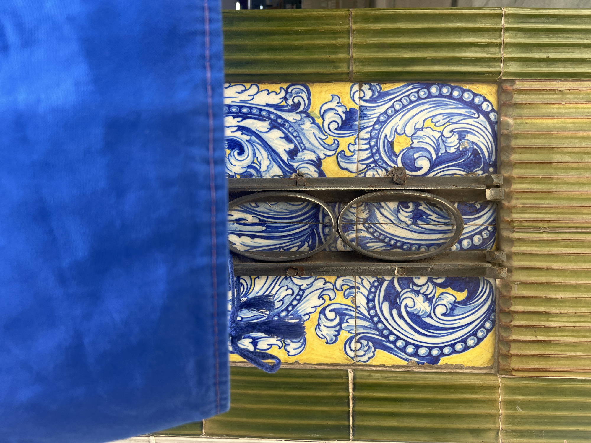 An exterior view captured from a low angle shows a decorative column in Barcelona, Spain. The column is adorned with a yellow tile featuring blue and white ornate patterns, set between vertical green ribbed tiles. A dark metal grate covers the central portion of the yellow tile, revealing two oval openings. A blue fabric drapes across the top of the image, obscuring the upper part of the column and featuring a blue tassel hanging from its edge. The lighting suggests an outdoor setting, possibly during the day.