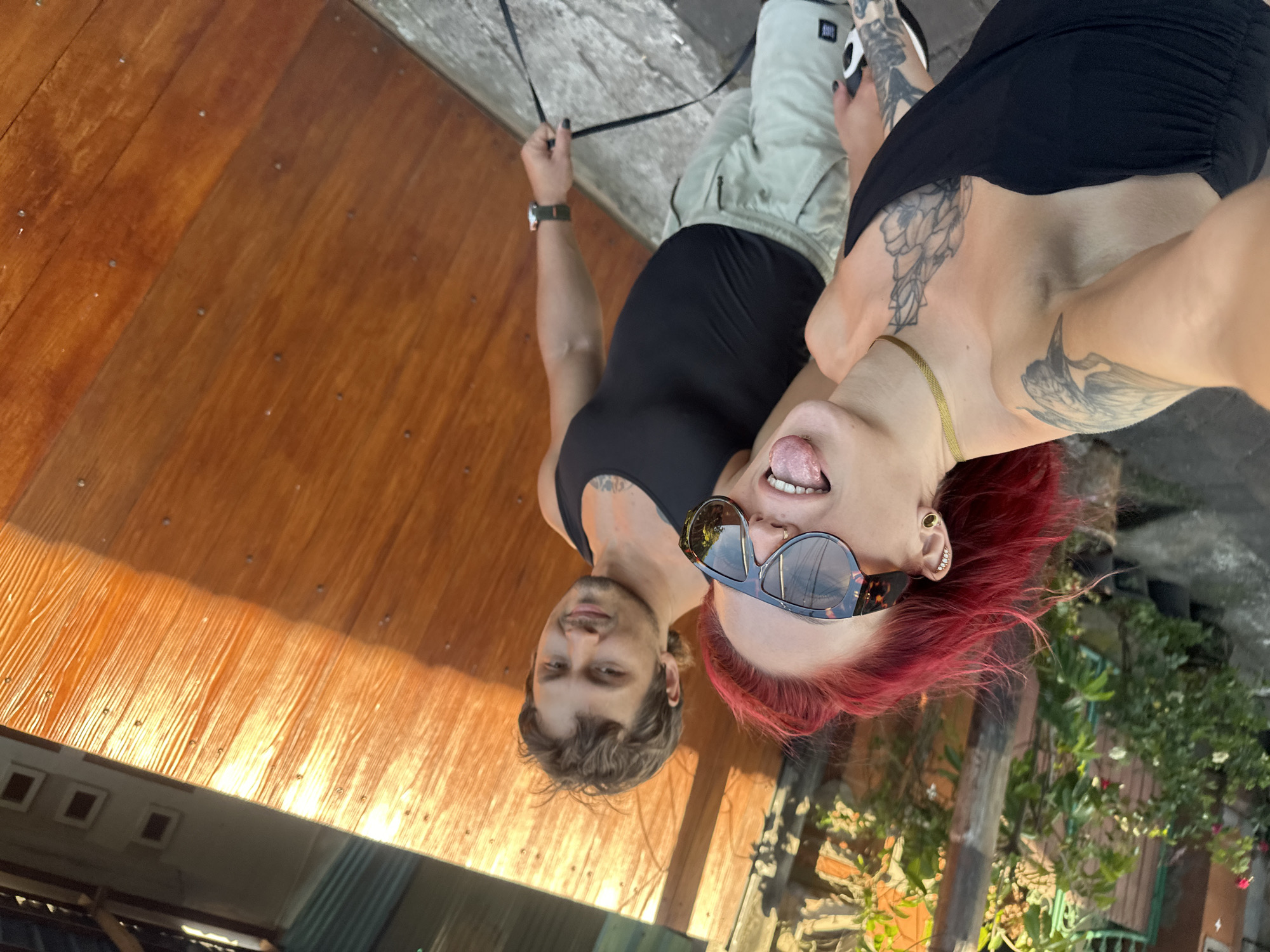 A selfie shows two individuals, a man and a woman, in an outdoor setting. The man, with short brown hair and light skin, is wearing a black tank top and a watch on his left wrist. The woman, positioned closer to the camera, has red hair, sunglasses, and multiple piercings on her left ear; she is sticking her tongue out and wearing a black top and a gold necklace. Both have tattoos visible on their upper bodies.
They are standing on what appears to be a wooden deck or platform, with a concrete wall visible in the background. Vegetation and a fence can be seen in the lower right corner. The location is likely North Kuta, Indonesia, indicated by the architectural style and flora. The perspective is skewed, suggesting the photo was taken at an angle, possibly using a selfie stick.