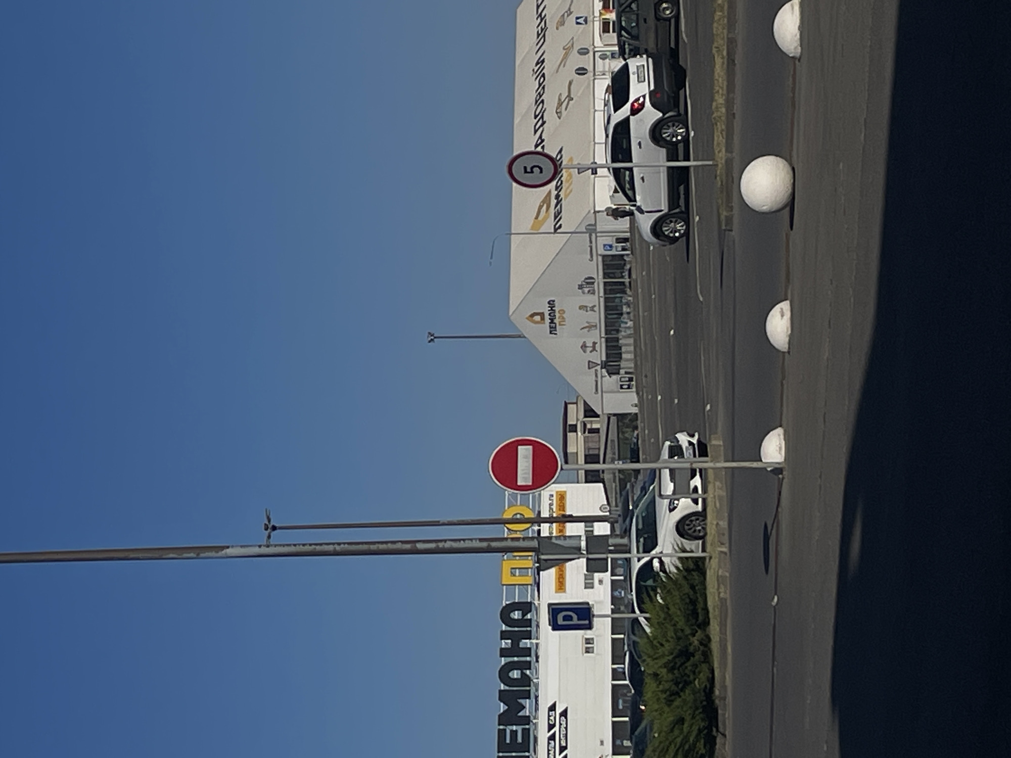 A view of a parking lot outside a large, white, tent-like structure with the word "ЛЕМАHA" visible on its side, indicating it's likely a marketplace or exhibition hall. A speed limit sign indicating "5" is present. Several cars are parked in the lot, including a white SUV and a white sedan. A "No Entry" sign is also visible. The sky is clear and blue. This scene is located in Verkhnetemernitsky, Russia.