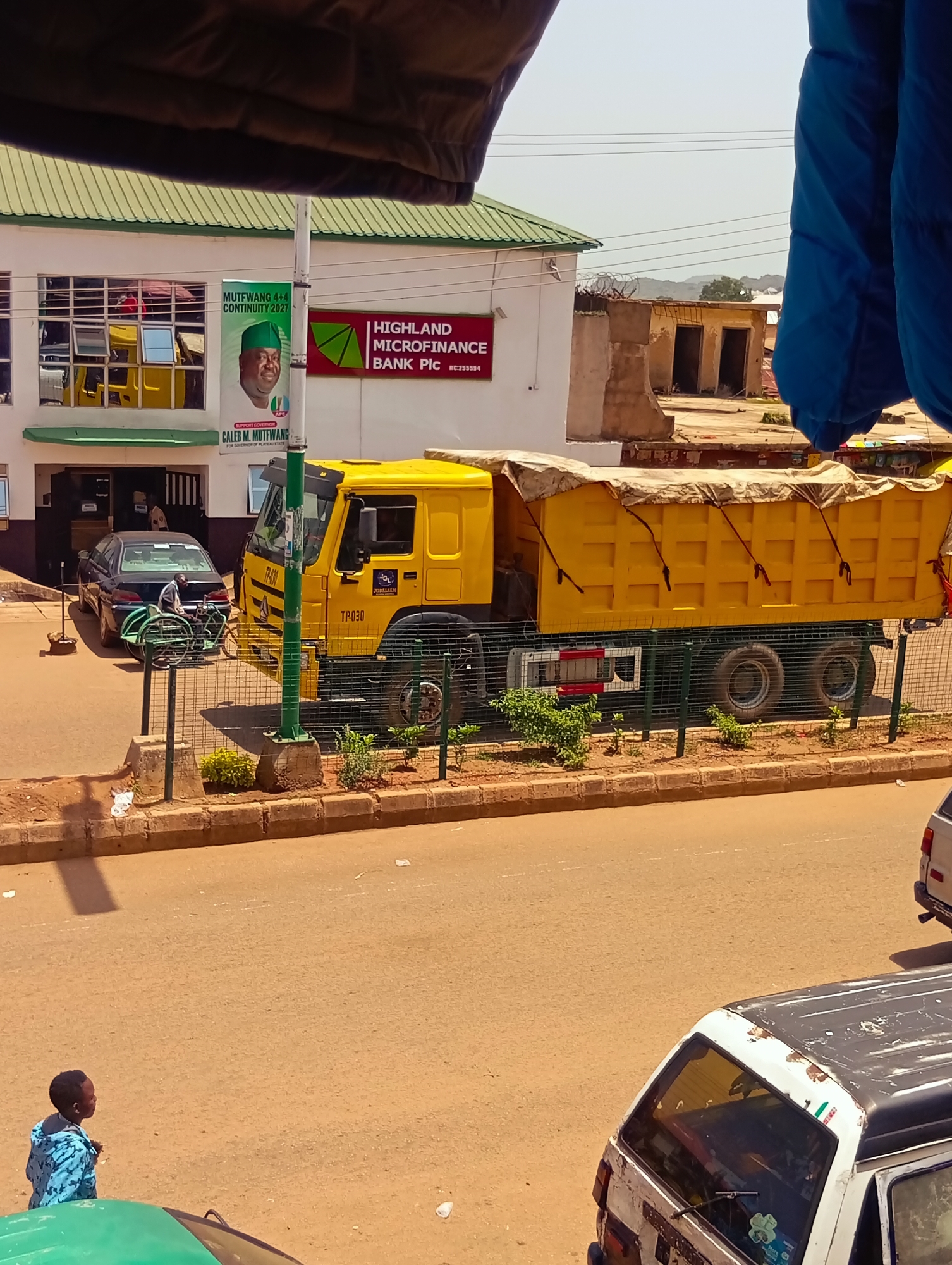 This image captures a street scene in Bukuru, Nigeria, under a bright, possibly midday sun, indicated by the strong shadows. The foreground is dominated by a yellow dump truck, its cargo covered with a beige tarp secured by ropes. The truck bears the inscription "TP-030" and a logo that includes "JODALEEM."
To the left of the truck is a building with the signage "HIGHLAND MICROFINANCE BANK Plc." Above the bank entrance, a poster features a man in a green cap, identified by text as "CALEB M. MUTFWANG," with slogans like "MUTFWANG 4+4 CONTINUITY 2027" and "SUPPORT GOVERNOR." A black sedan is parked in front of the bank, and a person is seated on a green bicycle rack behind it.
Further left, a young boy wearing a blue patterned shirt stands on the road, looking to his left. The street is paved and appears to be a busy thoroughfare, with other vehicles partially visible, including the roof of a white van in the lower right corner and a glimpse of a green vehicle in the lower left. The background shows more buildings and some vegetation, with hills visible in the distance, suggesting a somewhat urbanized area. The presence of the campaign poster points to a political event or election period. The overall atmosphere appears to be a typical day in a Nigerian town, with commerce and transportation in full swing.