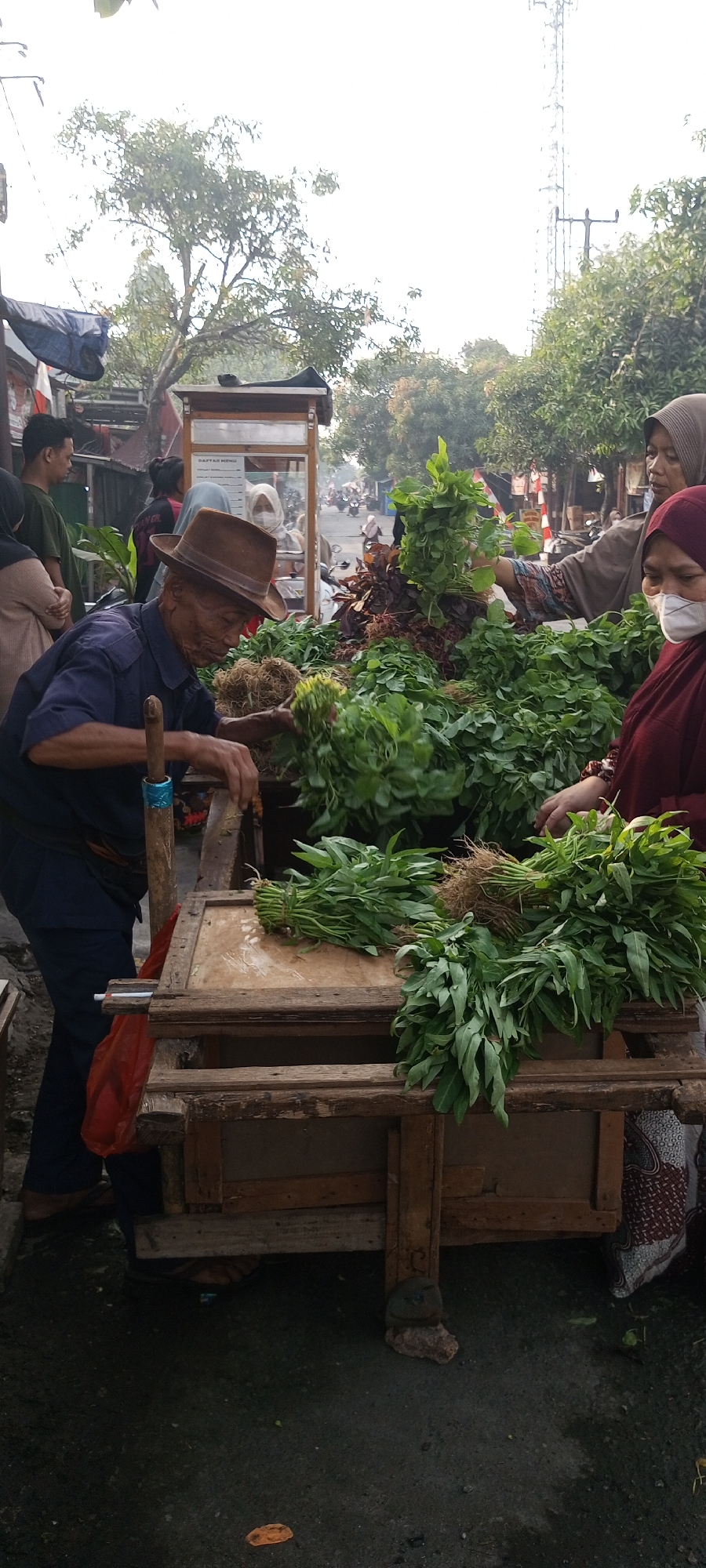 This image depicts an outdoor market scene in Setu, Indonesia, likely a street market. In the foreground, an elderly man wearing a brown fedora and a dark blue shirt is bent over a wooden cart filled with fresh green vegetables, appearing to be handling bundles of leafy greens. The cart has a wooden structure with a textured wooden surface. To his right, two women are interacting with the vegetables. One woman, wearing a brown hijab and a patterned garment, is reaching towards the vegetables. Another woman, wearing a burgundy hijab and a white face mask, is also visible. Behind the man and his cart, a small, covered food stall or kiosk is present, with a sign that reads "DAFTAR MENU". Several people are standing around, and in the background, there are trees, power lines, and a cell tower, along with a street with distant traffic including motorcycles. The ground is paved and appears slightly wet. The lighting suggests it is daytime, possibly morning or late afternoon, with overcast conditions.