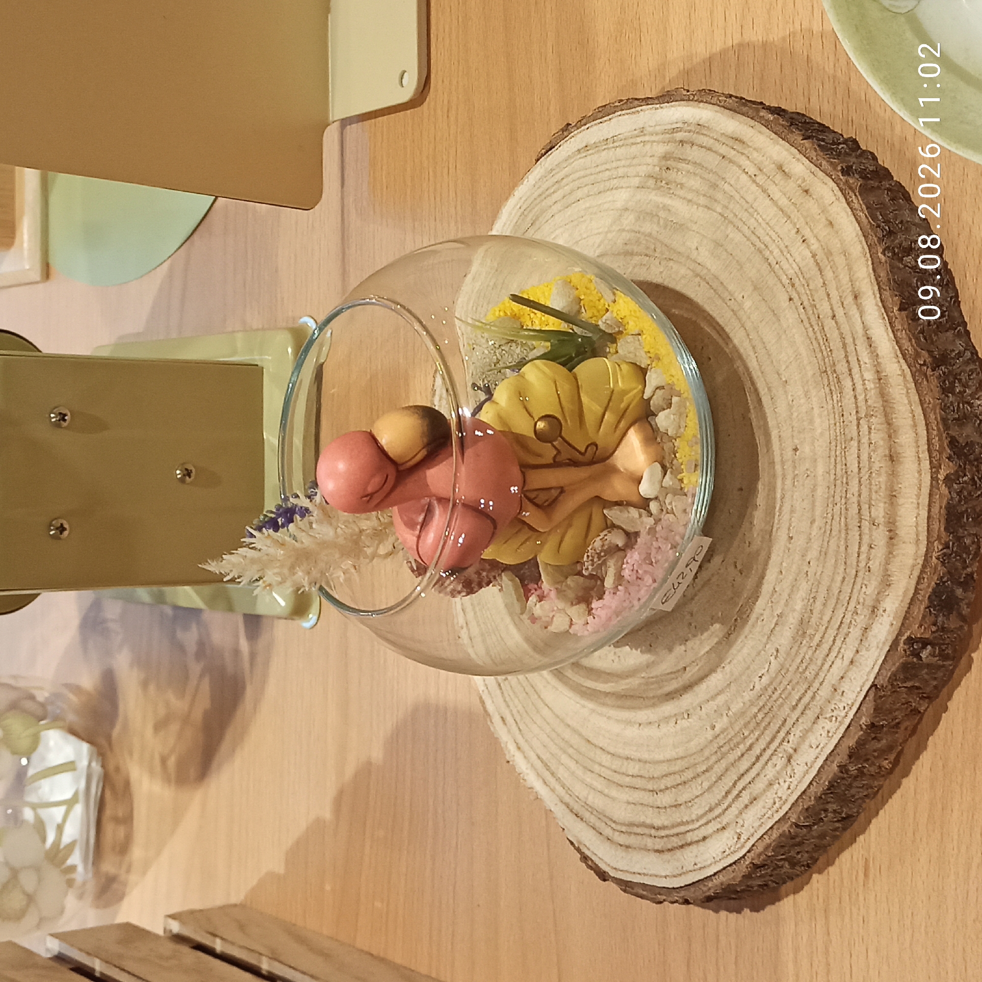 A decorative pink flamingo figurine sits in a clear glass bowl filled with pebbles, sand, and small shells. The bowl rests on a circular wood slice with visible rings. In the background, a beige, old-fashioned scale is visible, along with a sliver of a wooden crate and a pale green object. A timestamp "09.08.2026 11:02" is present in the lower right corner. The scene is set on a wooden table surface. No specific location in Molfetta, Italy, can be determined from the provided image.