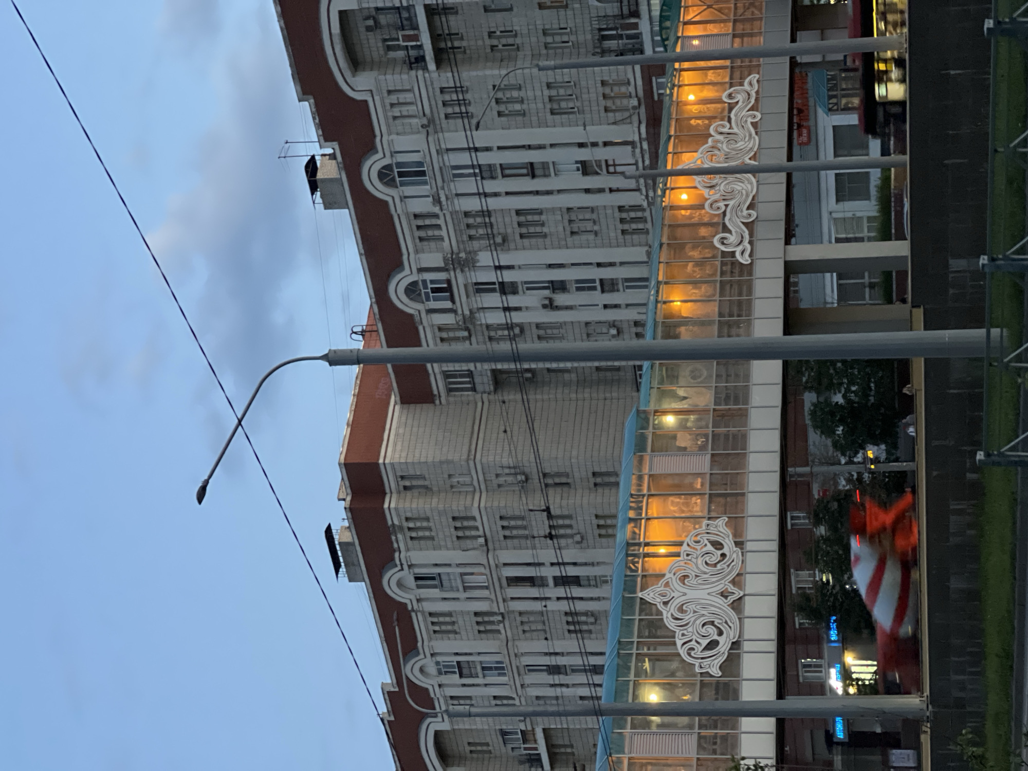An overpass with a glass enclosure and illuminated interior runs horizontally across the mid-ground of the image. The overpass features white decorative scrollwork on its exterior. Behind the overpass, a large, light-colored apartment building with a red roof and arched windows dominates the upper portion of the frame. To the left of the apartment building, a street lamp on a pole is visible, and power lines crisscross the sky. In the foreground, a blurred red and white object, possibly a construction vehicle or traffic cone, is moving from left to right. The sky is a dusky blue with some clouds. The scene appears to be urban, potentially in Kazan, Russia, given the architectural style and the presence of public transport like buses visible in the background.
