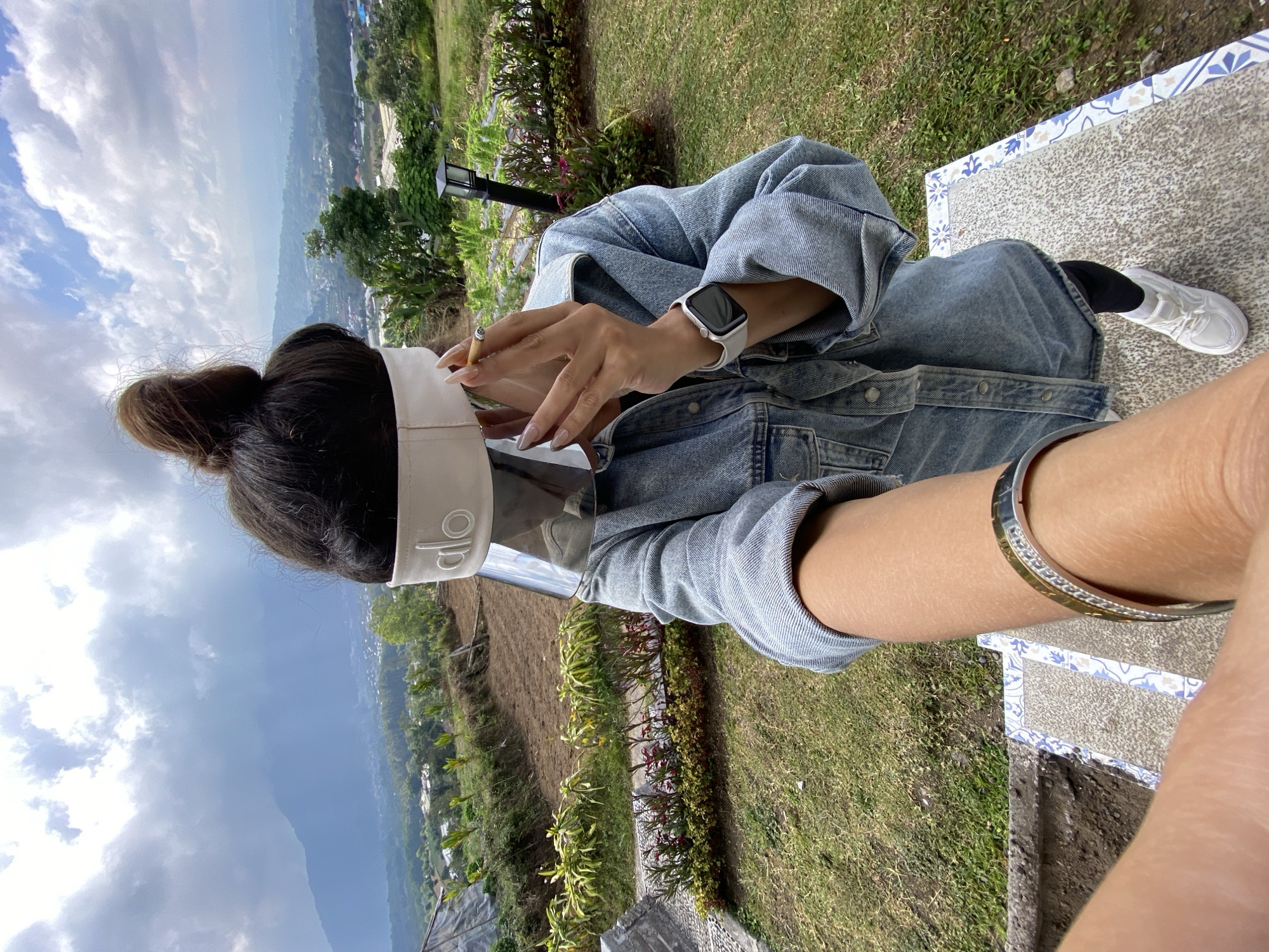 A person stands on a patterned staircase outdoors, holding a lit cigarette to their lips. They are wearing a denim jacket, black leggings, and white sneakers, with a face shield and a white headband with the "alo" logo visible. A smartwatch is on their left wrist, and a bracelet adorns their right wrist. The background reveals a lush, green landscape with terraced fields and distant mountains under a cloudy sky, consistent with the scenery around the Ulun Danu Beratan Temple in Baturiti, Bali, Indonesia.