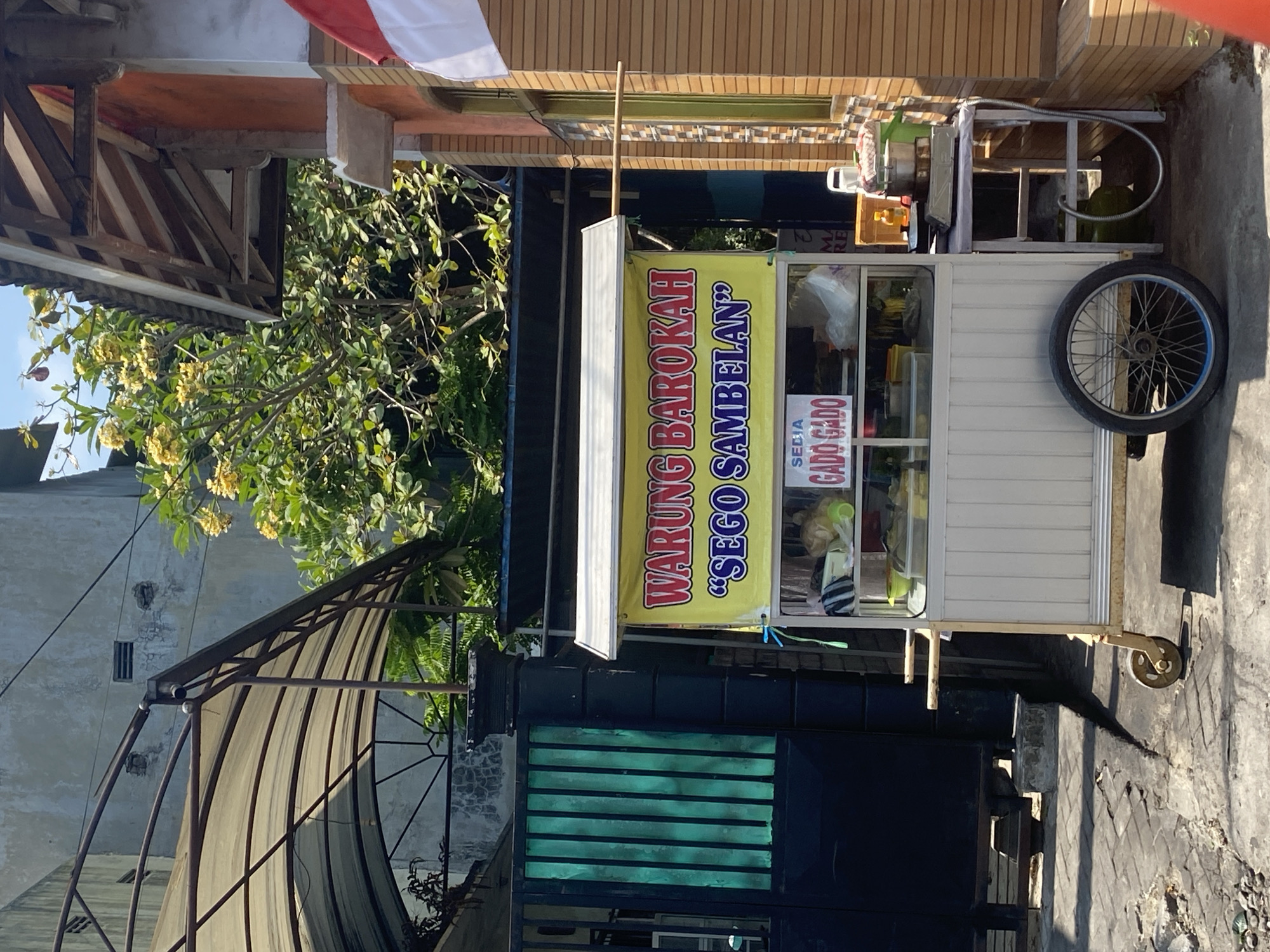 The image shows a street-side food stall in Kediri City, Indonesia, on a sunny day. The stall, named "WARUNG BAROKAH SEGO SAMBELAN," is a mobile cart with a white body and yellow awning. It is positioned on a paved walkway next to a building with brown wooden siding and an orange-colored section. A partially visible Indonesian flag is attached to the building.
In front of the stall, a sign advertises "SEGO GADO GADO." The cart itself contains shelves displaying various food items and containers. A small cooking stove and a gas cylinder are visible on a table next to the stall. The background includes a black metal gate with green vertical slats, a large tree with yellow flowers, and a building with a gray, weathered facade. The ground is a rough, gray paved surface, with some debris scattered around. The scene appears to be a typical urban environment in Indonesia, likely a commercial or residential street. There are no people visible in the image.