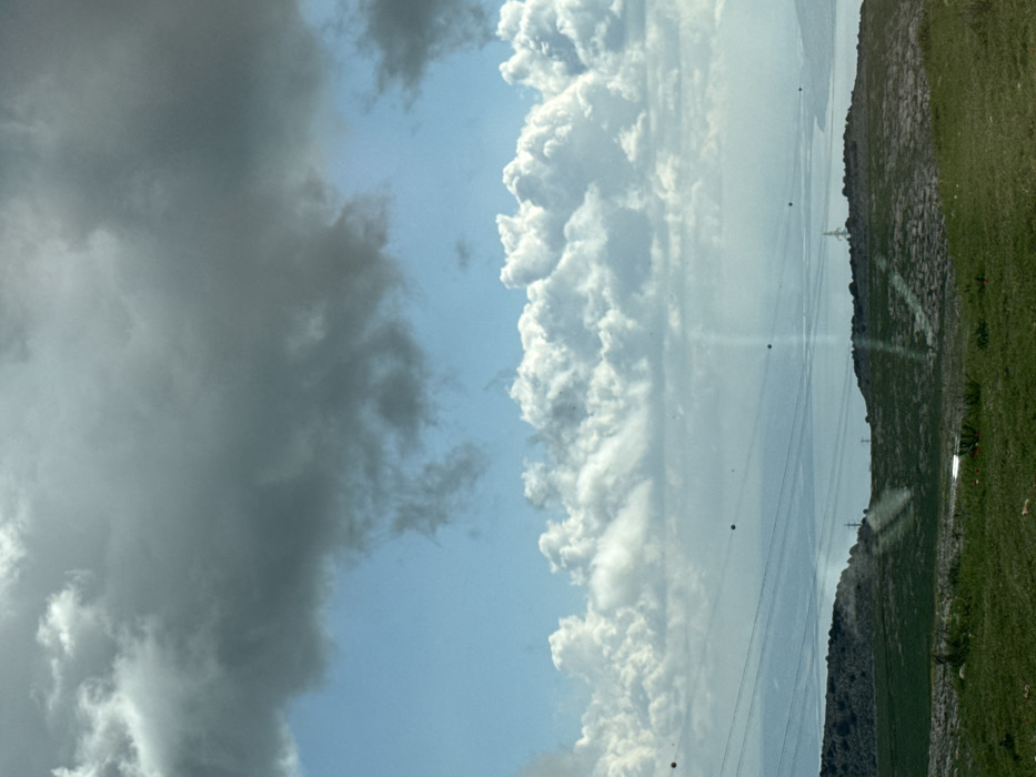The image displays a panoramic landscape view, likely captured from an elevated position overlooking the Sea of Galilee (Lake Kinneret) and surrounding terrain in the city of Safed, Israel. The sky occupies the upper two-thirds of the frame, characterized by a dynamic cloud formation: dense, dark gray stratocumulus clouds dominate the uppermost portion, transitioning to bright white cumulus clouds at mid-sky, with patches of clear blue sky visible between the layers.
Across the mid-ground, a broad expanse of water, consistent with the Sea of Galilee, extends horizontally, appearing blue and somewhat hazy with visible light reflections on its surface. The water is bordered by undulating, green hills that recede into the distance. In the foreground and closer mid-ground, the terrain is characterized by rolling hills covered predominantly in green grass, with some areas of sparse vegetation and exposed earth. Small red flora, possibly poppies, are visible in the immediate grassy foreground.
Several parallel power lines equipped with spherical, dark-colored aviation markers (likely orange or black) traverse the scene horizontally across the lower sky and above the hills, extending from the left to the right side of the frame. One prominent metal electricity pylon is visible on a hill to the right, and another partial pylon is discernible further left. The scene is illuminated by sunlight, creating varying degrees of brightness and shadows across the landscape. No human figures or active subjects are present. The viewpoint appears to be from within a vehicle or behind glass, as evidenced by subtle light flares or reflections in the lower half of the image.