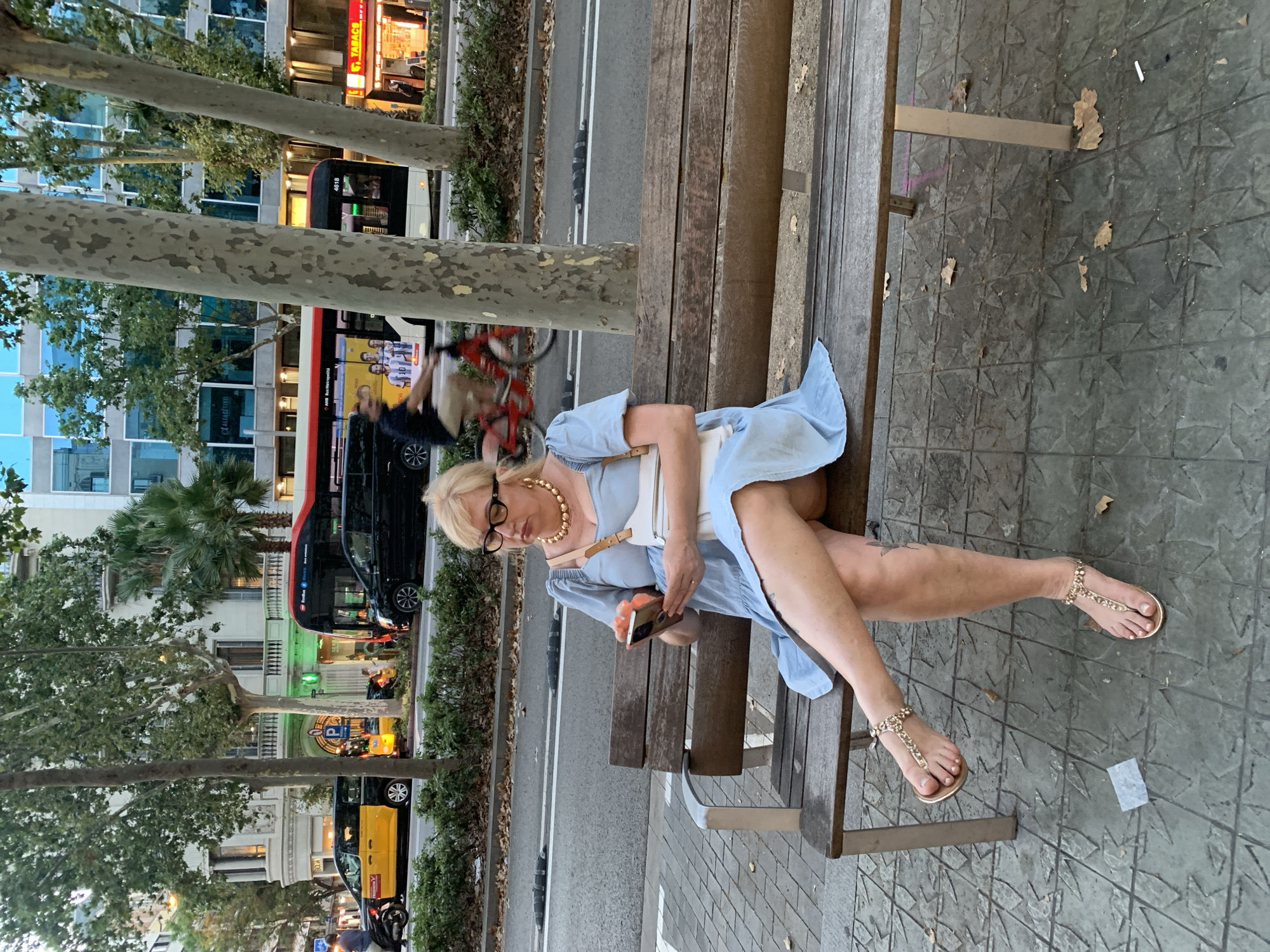 A middle-aged Caucasian woman with blonde hair and glasses is sitting on a wooden bench, looking at her smartphone. She is wearing a light blue dress, a large gold necklace, and gold sandals. A white purse is on her lap. She is sitting on a tiled sidewalk next to a street with a yellow and black taxi and a bus. The scene appears to be in Barcelona, Spain, given the distinctive architecture and the presence of the yellow taxi, which is a common sight in Barcelona. The street is lined with trees and buildings.