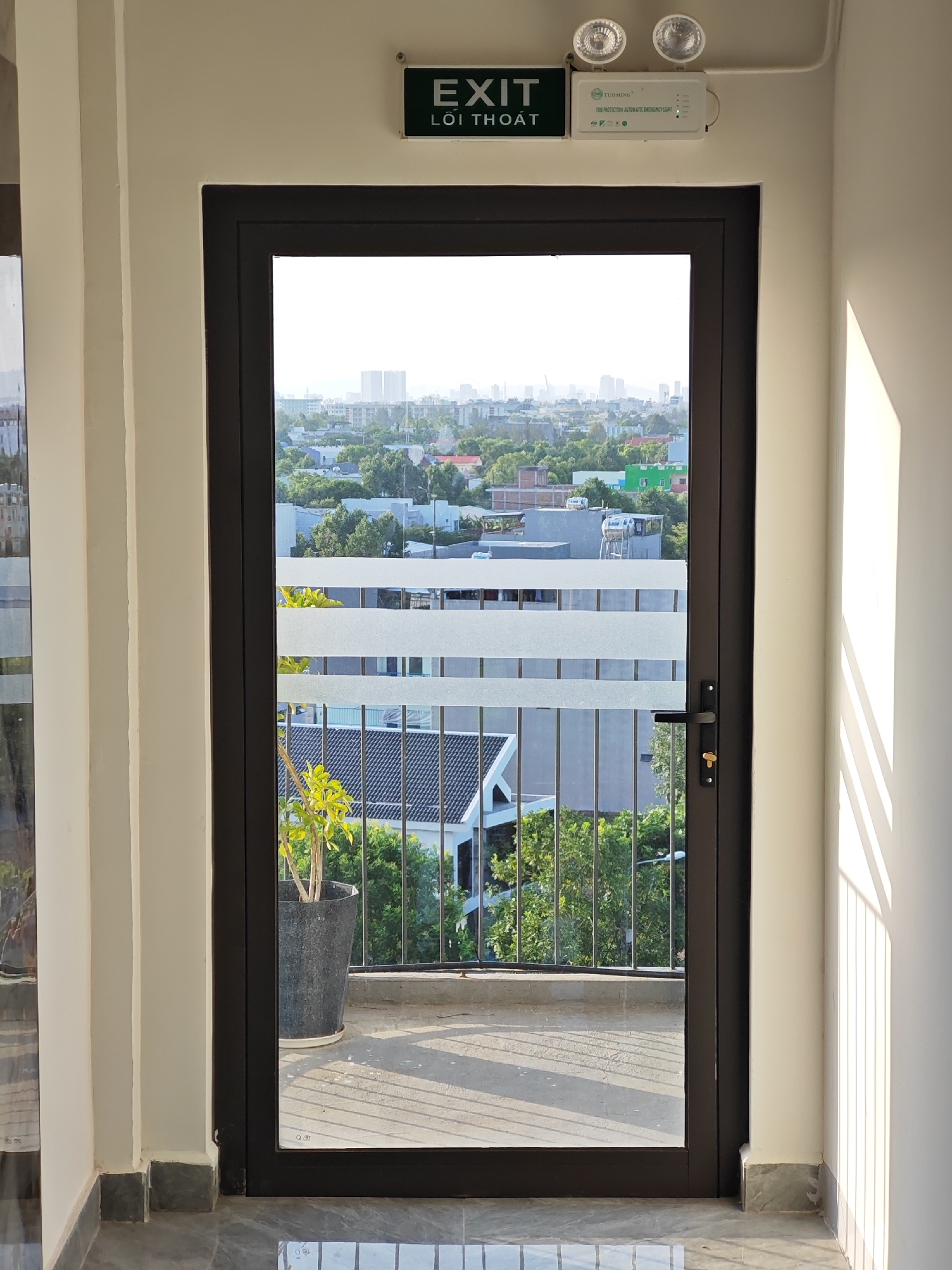 A dark-framed glass door opens to a balcony overlooking a city skyline. Above the door, a green EXIT sign with Vietnamese text is illuminated, alongside an emergency light fixture. The balcony features a potted plant and a metal railing. The sun casts long shadows across the concrete floor, indicating a late afternoon or early morning. The city view includes buildings and lush green trees, suggesting a location in a developed urban area of Da Nang, Vietnam.