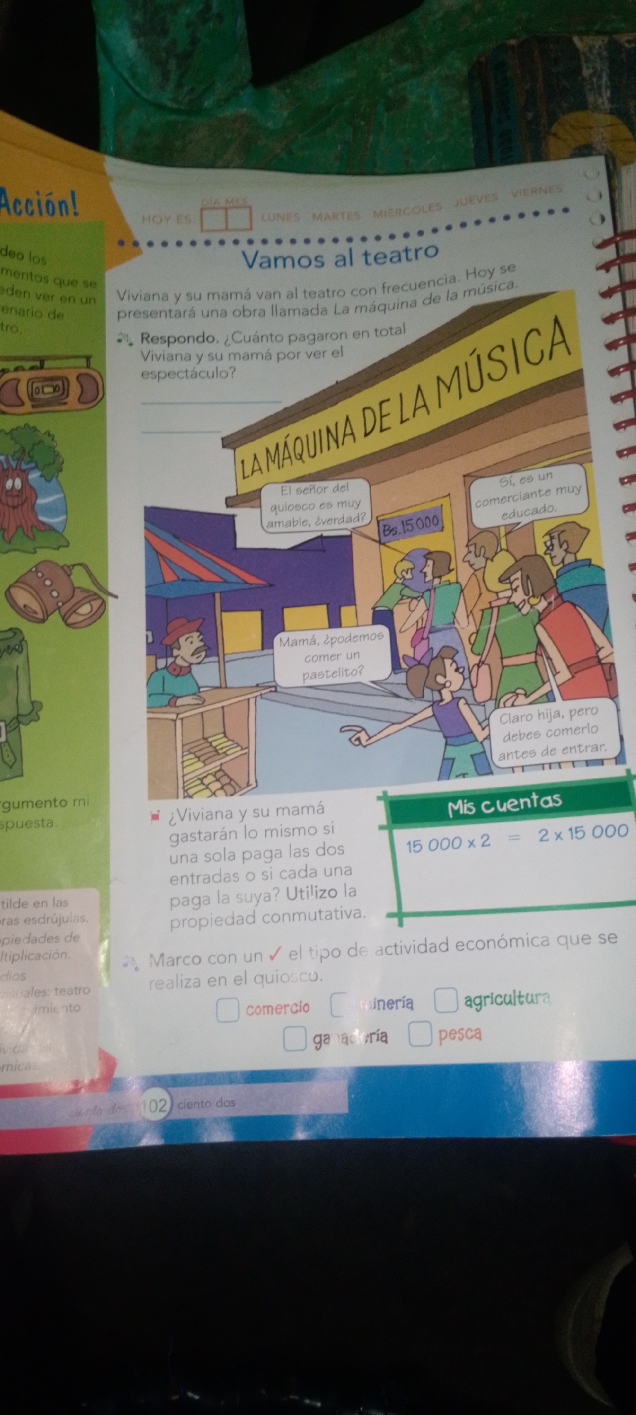 The image is a close-up, overhead shot of an open workbook page, likely from a child's educational material, featuring a colorful illustration and text. The illustration depicts a busy scene outside a building labeled "LA MÁQUINA DE LA MÚSICA" (The Music Machine). A group of people, some queuing, are interacting in front of the building. A child is asking a woman, presumably her mother, if they can eat a pastry. The woman is responding that they must eat it before entering. Another speech bubble indicates that the kiosk vendor is very kind and educated, and a price of "Bs. 15.000" is visible. The workbook page includes text related to a story about Viviana and her mother going to the theater and questions about the cost of tickets. A section titled "Mis Cuentas" (My Accounts) shows a multiplication problem: "15 000 x 2 = 2 x 15 000", illustrating the commutative property of multiplication. There is also a prompt asking to mark the type of economic activity related to a kiosk, with options including "comercio" (commerce), "minería" (mining), "agricultura" (agriculture), "ganadería" (livestock), and "pesca" (fishing). The page is well-lit, showing the details of the illustration and text clearly. The background of the image is dark, suggesting the workbook is lying on a dark surface. There is no discernible information within the image that indicates a location in La Tagua, Venezuela.