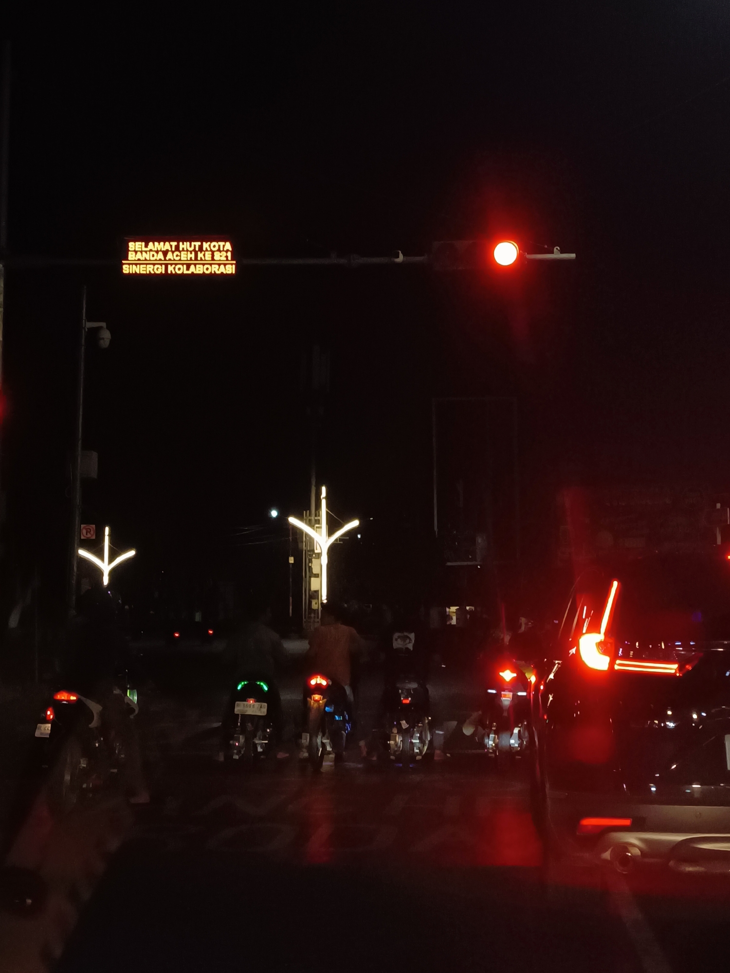 The image shows a street scene at night in Banda Aceh, Indonesia. A digital billboard displays the text "SELAMAT HUT KOTA BANDA ACEH KE 821 SINERGI KOLABORASI" in yellow, which translates to "Happy 821st Anniversary of Banda Aceh City, Synergy Collaboration." A red traffic light is visible overhead, indicating a stop. Several motorcycles are present, some with illuminated license plates and decorative lights. A dark-colored vehicle with illuminated taillights is in the foreground on the right. Streetlights with decorative designs are also visible. The overall environment appears to be a city street at night with moderate traffic.