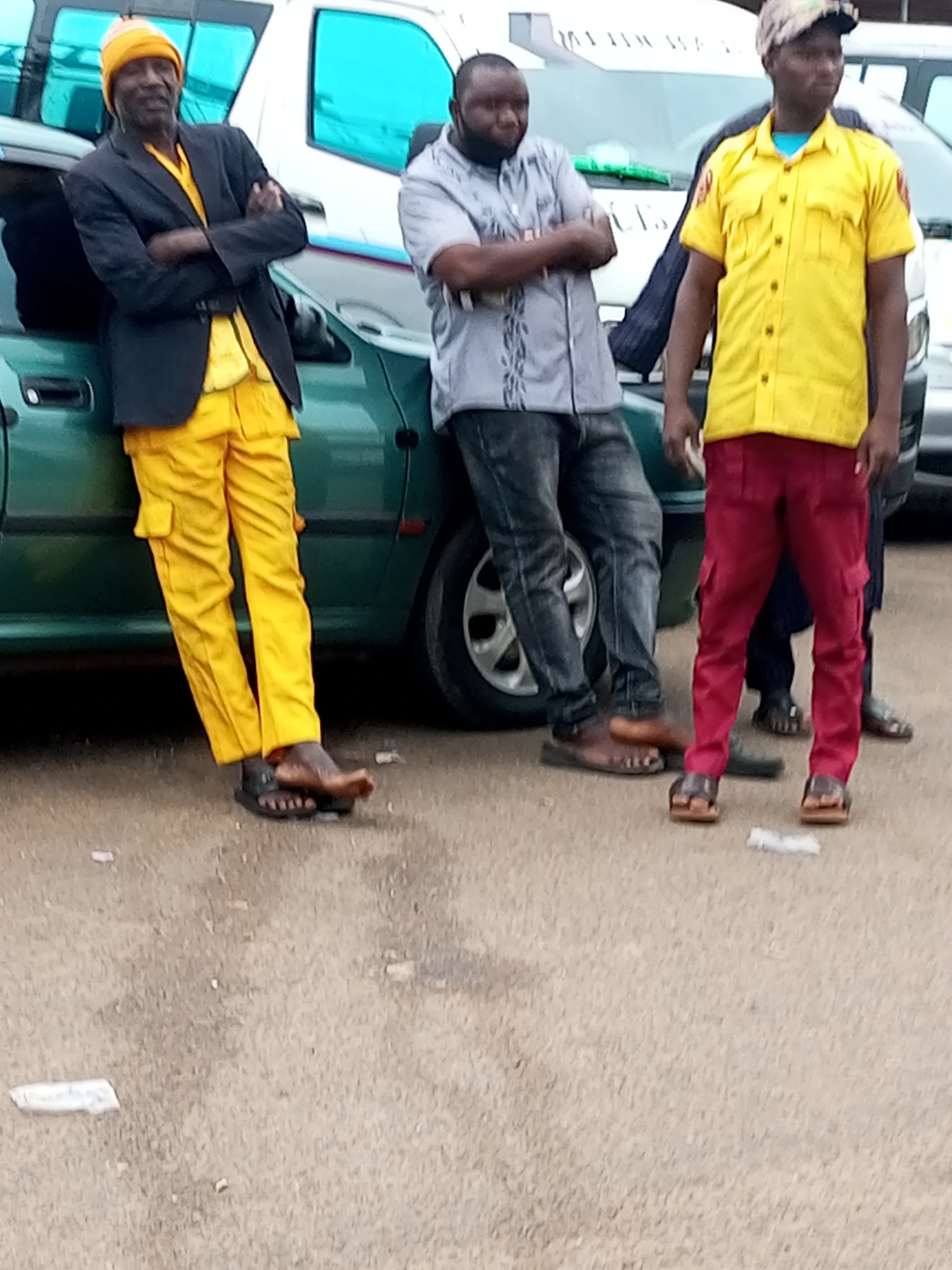 In Gombe, Nigeria, on a dusty, paved surface, three men stand near a green sedan. The man on the left, wearing a yellow beanie, a black blazer over a yellow shirt and yellow cargo pants, has his arms crossed. To his right, a man in a grey shirt and jeans leans against the car. The third man, on the far right, is dressed in a yellow shirt and red cargo pants. The background shows the side of a white vehicle with tinted windows.