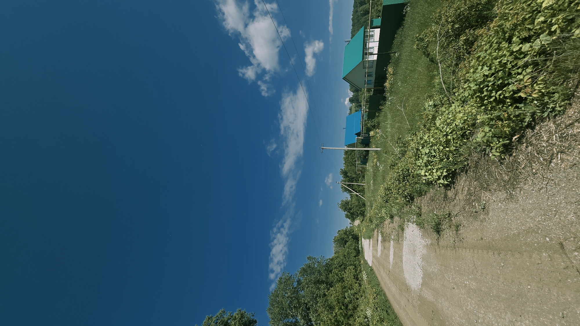 The image depicts a rural scene in Kzyl-Chishma, Russia, under a clear, deep blue sky with scattered white clouds. A dirt road, marked by several water puddles, winds its way from the foreground into the distance, bordered by lush green vegetation and trees. On the right side of the road, a row of houses with distinctive bright blue and green roofs is visible. A green fence runs along the property line of these houses. Electrical poles and power lines are also present in the scene, stretching across the landscape. The overall impression is of a tranquil, sunlit day in a countryside setting.