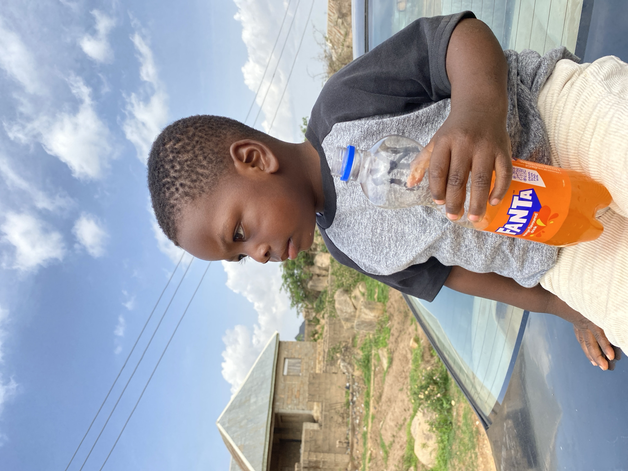 A young Black boy stands outdoors in Barakin Akawo, Nigeria, holding a bottle of Fanta. He wears a two-toned shirt, dark sleeves and grey torso, and appears to be near a black car. In the background, there is a partially constructed building and a clear sky with scattered clouds. Power lines are visible in the upper left corner.