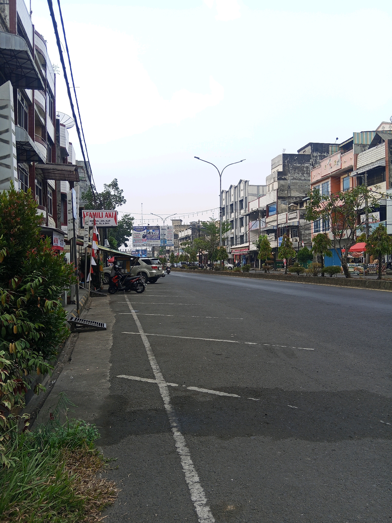 The image depicts a street scene in Lubuklinggau, Indonesia, likely on Jalan Yos Sudarso. On the left side of the street, a row of multi-story buildings with shops on the ground floor lines the sidewalk. Some buildings have awnings and balconies, and satellite dishes are visible on some rooftops. A prominent sign reads "FAMILI AKI" in red and white. A lush green bush is in the foreground on the left. The sidewalk area has a drain cover.
The street itself is paved and appears to have multiple lanes. White parking lines are visible on the asphalt. Several vehicles are parked along the street, including a motorcycle and an SUV. A few trees are planted along the median or sidewalk. In the background, more buildings and streetlights are visible, along with several large billboards and advertisements. The sky is overcast and appears bright. The overall scene suggests a typical commercial street in an Indonesian city during daytime.