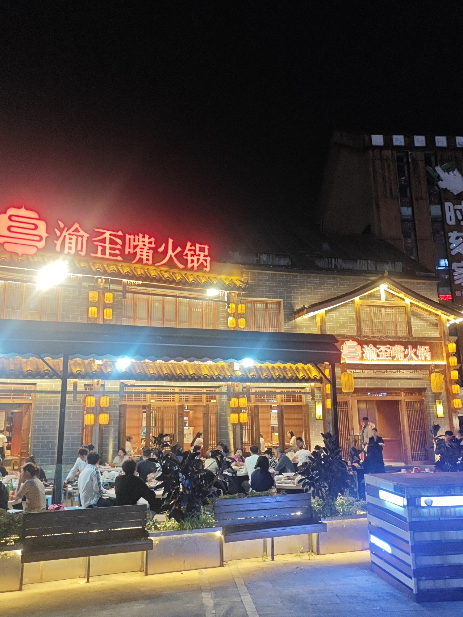 The image depicts an outdoor dining area at night in Chongqing High-tech Zone, China. The focal point is a restaurant with traditional Chinese architecture, featuring a sign in red neon lights that reads "渝歪歪嘴火锅" (Yú wāi wāi zuǐ huǒguō), indicating a hot pot restaurant. The restaurant has a multi-tiered roof, gray brick facade, and numerous lit yellow lanterns. People are seated at tables, presumably dining. There are benches and planters in the foreground, and a modern building is visible in the background to the right. The overall ambiance is illuminated and suggests a vibrant, active dining scene.