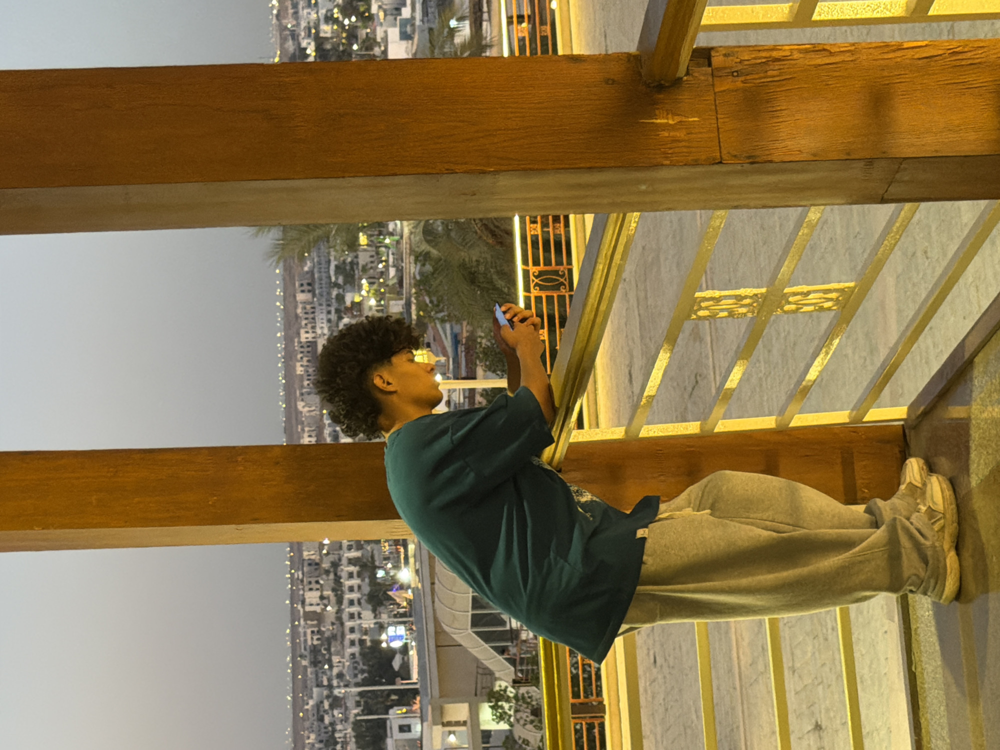 A young person with curly dark hair is standing on a balcony overlooking a city at dusk. They are wearing a dark teal t-shirt and light gray sweatpants. The person is leaning on a yellow railing, looking down at their mobile phone, which they are holding in their hands. The background shows a cityscape with many buildings and lights, indicating a populated area. Palm trees are visible in the middle ground. The balcony itself is made of wood and metal railings, with a tiled floor. The overall lighting suggests it is either early morning or late evening. The location is identified as Sharm El Sheikh, Egypt.