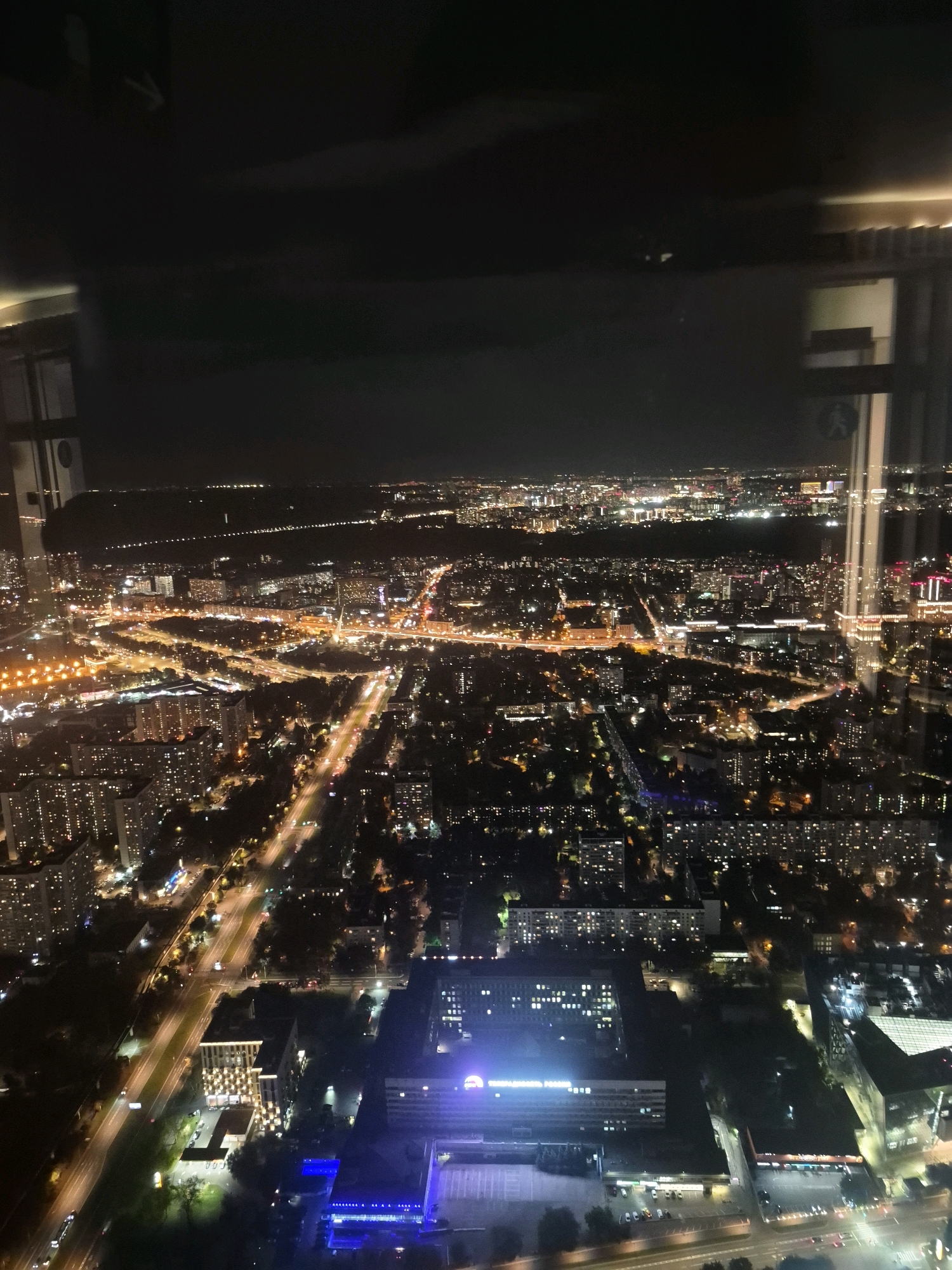 This is an aerial nighttime photograph of a city skyline. The image captures a dense urban landscape with numerous buildings illuminated by artificial lights, indicative of a city at night. Roads crisscross the scene, with car headlights and taillights creating streaks of light, suggesting active traffic. The perspective is from a high vantage point, possibly a tall building or an aircraft, offering a panoramic view of the city. In the foreground and midground, apartment buildings with many lit windows are visible, as well as larger commercial or public buildings. The background extends to a horizon dotted with more distant lights, suggesting the vastness of the urban sprawl. Hints of greenery, likely trees, can be observed interspersed between the buildings, particularly in the darker, less developed areas. The overall impression is one of a bustling metropolis after dark.
Based on visual cues such as the architectural style of some buildings and the layout of the road networks, this scene is likely in or around Khimki, Russia. Specifically, the prominent tall building visible in the mid-right portion of the image, with its distinctive illumination, could be a landmark. The general density of residential buildings and the multi-lane highway infrastructure are consistent with urban areas in Russia. Without specific identifying features like unique landmarks or signage that are clearly legible at this resolution, pinpointing the exact location within Khimki is difficult, but the overall urban character strongly suggests a Russian city.