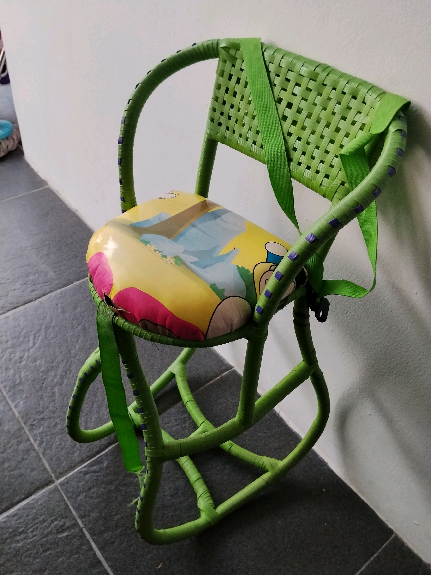 A green rattan child's chair with a cartoon-themed seat is positioned against a white wall on a tiled floor. The chair is leaning slightly, with its legs splayed. A green strap is attached to the back of the chair, extending down and to the side. The seat of the chair is yellow with pink and blue cartoon graphics. There is a small tear on the left side of the seat. The floor is made of dark grey tiles. The lighting suggests an indoor environment, likely a home or a covered outdoor space in Sukabakti, Indonesia.