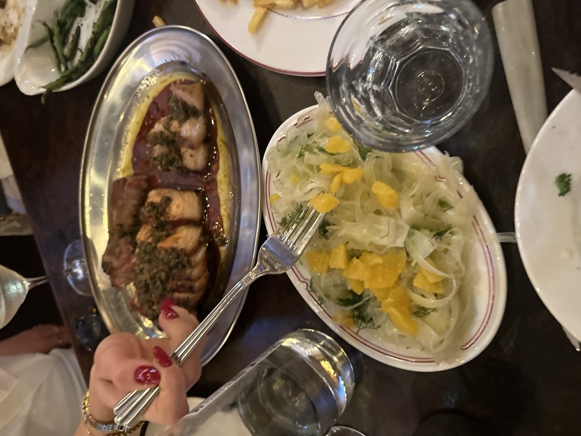 A close-up shot shows a table setting with food and drinks. In the foreground, a small white plate holds a salad with thinly sliced white vegetables, green herbs, and orange pieces. A silver fork is partially submerged in the salad. To the right, a clear glass with a textured surface is filled with water. In the background, a large silver platter contains a dish of meat with a dark sauce and green toppings, and a white bowl filled with green beans. A person's hand with bright red nail polish is visible on the left, holding a set of silver utensils. The overall impression is of a meal being served in a restaurant, with the dark wood grain of the table visible.