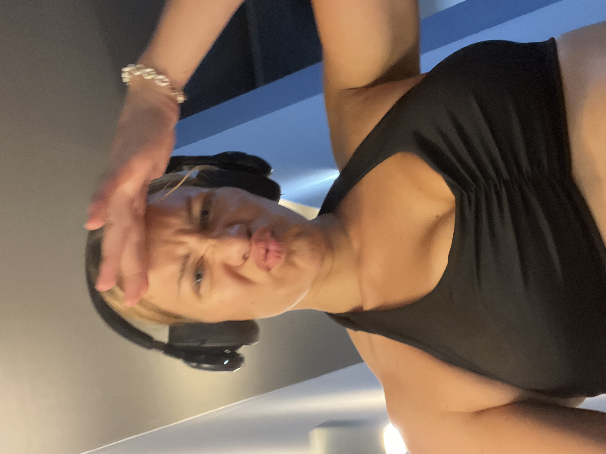 A woman wearing headphones and a black sports bra is captured in a close-up, low-angle shot. She has her right hand raised to her forehead, shielding her eyes, and is making a pouting face. The background is blurred, showing a hint of blue and grey. The image was taken in a gym in Altötting, Germany.