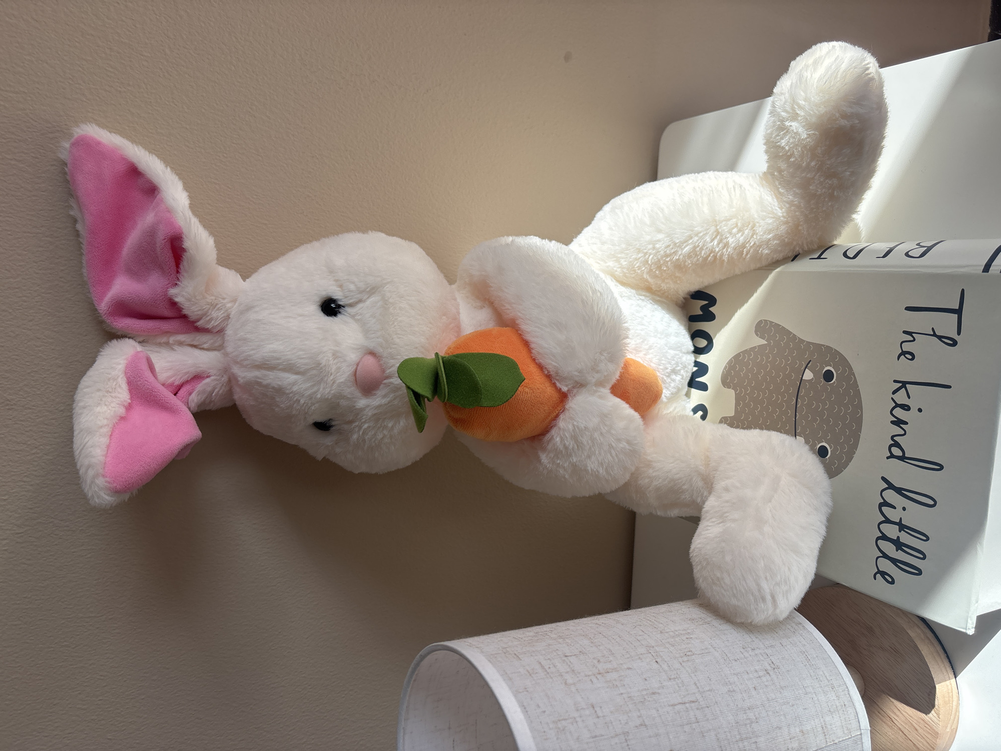 An indoor still life composition is captured in this medium shot photograph. The primary subject is a plush white bunny toy, approximately 18 inches in height, seated on a stack of two books. The bunny is positioned centrally in the frame and is facing slightly to the left. It has pink inner ears and a pink nose. The bunny is holding a small orange plush carrot with green fabric leaves. The bunny's front paws are clasped around the carrot, and its legs are extended forward.
The bunny is seated on two books that are stacked horizontally. The top book appears to be a children's book with illustrations of a stylized beige hippo-like creature and text that reads "kind little" in dark blue script, and "NOW" in dark blue sans-serif font. The book below it is mostly obscured, but some dark blue text is visible on its spine.
To the left of the bunny and books, a portion of a white lampshade with a textured surface and a wooden base is visible. The background consists of a plain, light beige wall with subtle shadows cast by overhead lighting, possibly sunlight. The scene is illuminated by light coming from the right, creating soft shadows on the wall and highlighting the textures of the bunny and the lampshade.
The photograph is taken from a slightly elevated viewpoint, looking down at the scene. The overall impression is one of warmth and domesticity. There is no indication of the city of Dubai, United Arab Emirates, in the image.
