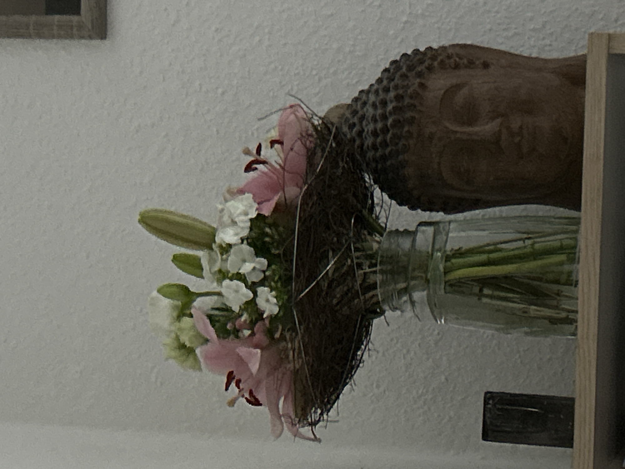 A close-up, eye-level shot shows a bouquet of pink and white lilies in a glass vase. The bouquet is nestled in a dark, woven material that resembles a bird's nest. To the right of the vase, a brown stone Buddha head is visible. The background is a textured, off-white wall. A dark, rectangular object rests on a wooden shelf to the left of the vase. The scene appears to be an indoor still life, possibly in a residential setting in Waldbröl, Germany, given the context of the request.