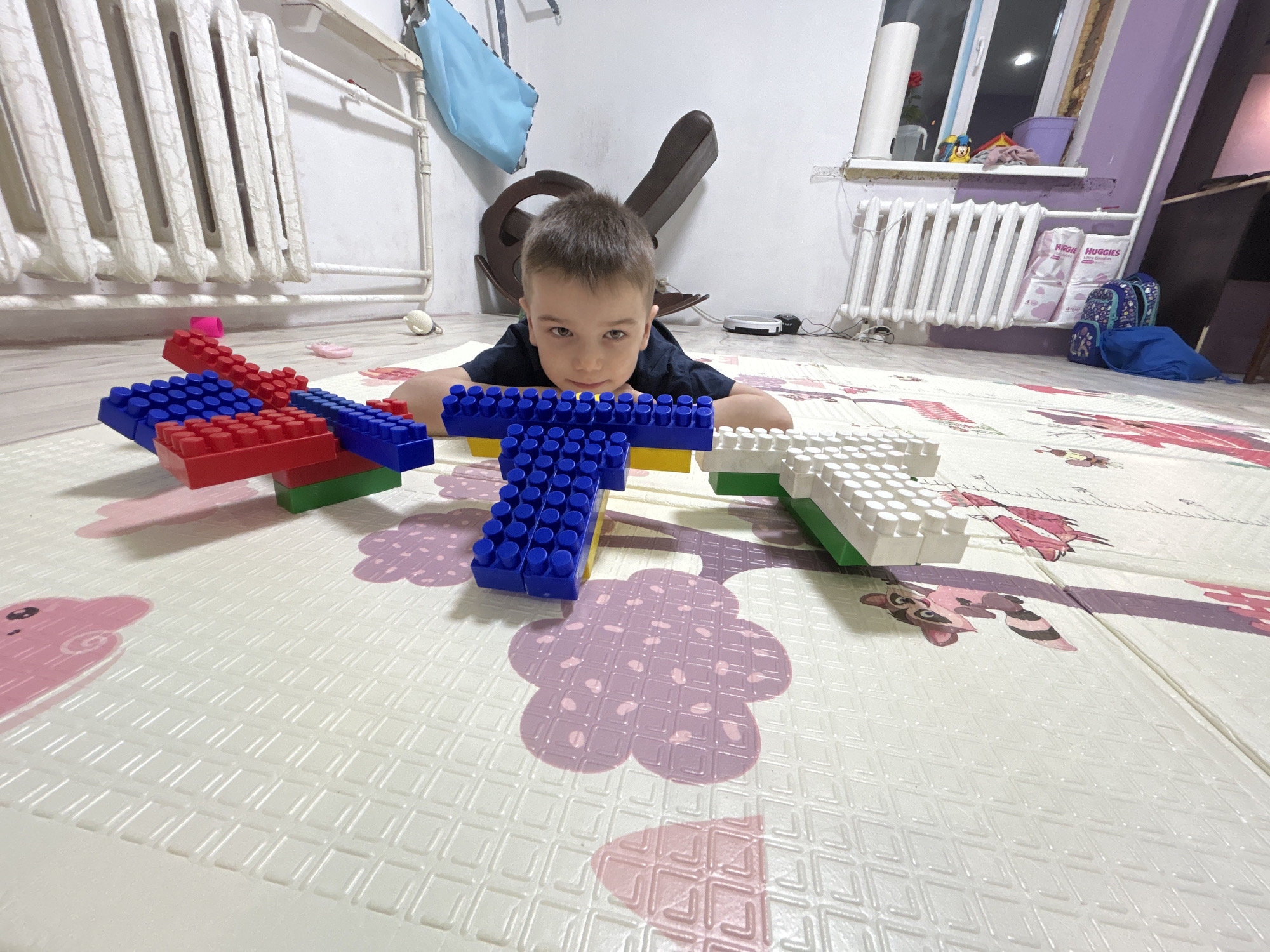 A young boy lies on a white and pastel colored play mat in what appears to be an indoor setting in Rusalevka, Russia. The mat is patterned with images of clouds, flowers, and raccoons. The boy has short brown hair and is wearing a dark-colored t-shirt. He rests his arms on a formation of large plastic building blocks arranged in front of him. The blocks are various colors: red, blue, yellow, green, and white. Behind the boy are a white radiator, a blue cloth hanging from a rack, and a dark-colored rocking chair. To the right are another white radiator, bags of Huggies diapers, and a blue bag. A windowsill with items on it is visible above the radiator, with purple walls in the background. A dark shelf unit is also visible on the right.