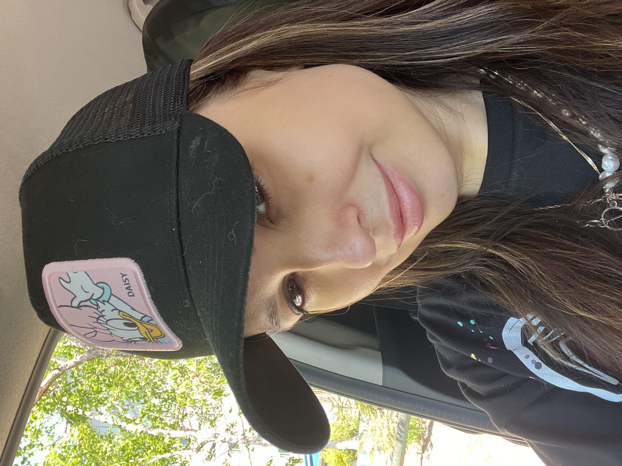A selfie of a woman with long brown hair wearing a black trucker hat with a Daisy Duck patch. She is wearing a black shirt with a white graphic design. She appears to be inside a car, with the interior visible. Outside the car, green foliage and possibly a building are visible. The location is unknown.