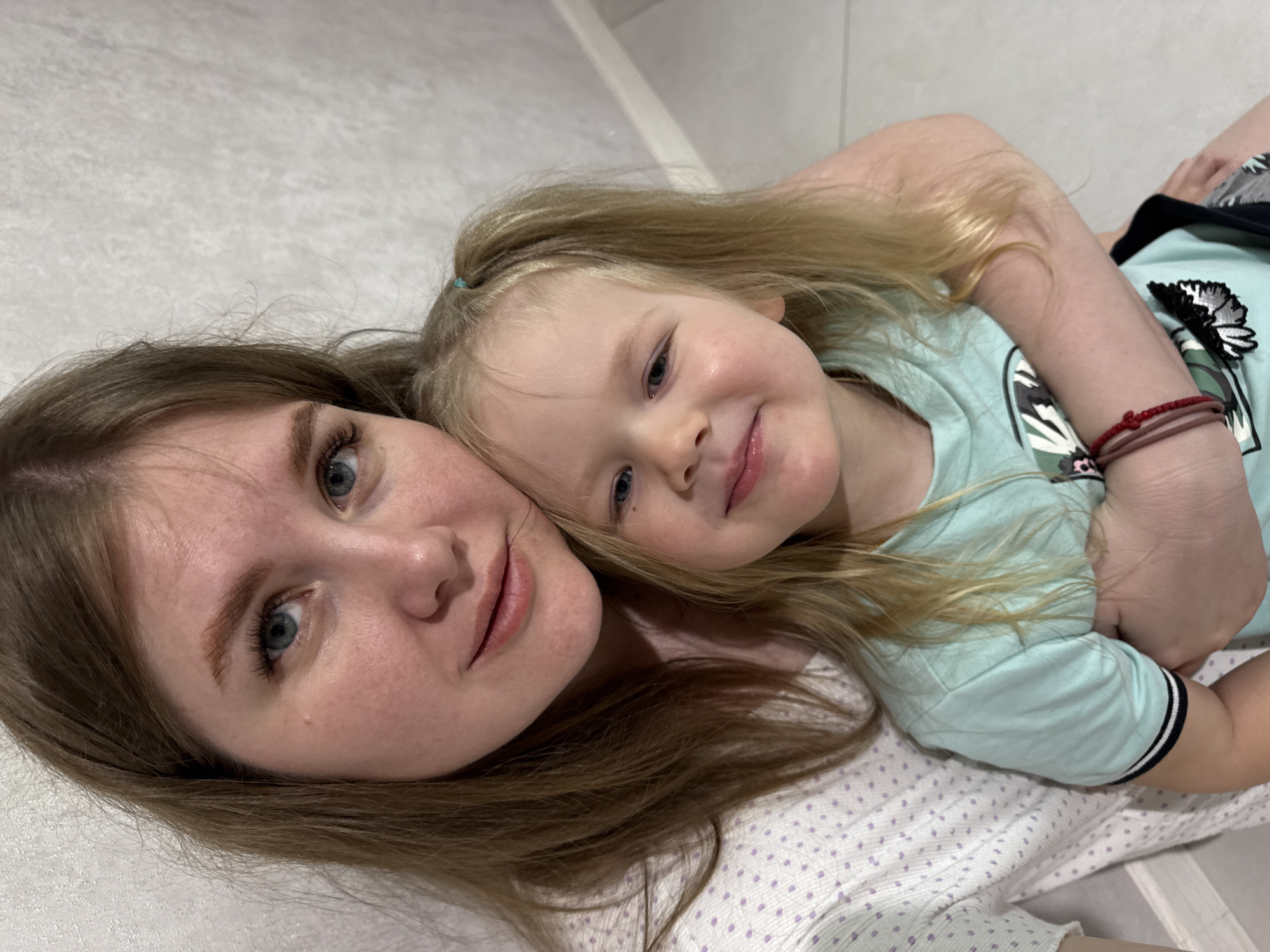 In Gornyi (Dvurechenskoe S/P), Russia, an image captures an adult female embracing a young female child indoors. The adult female, positioned to the left, has light brown hair and blue eyes, wearing a white short-sleeved top with purple polka dots. She directs her gaze towards the camera with a slight smile, with her left arm around the child. The child, a young female, positioned in the foreground, has blonde hair styled in two pigtails secured with light blue ties, and blue eyes. She wears a light teal short-sleeved t-shirt with a partial graphic design and a red string bracelet on her left wrist. Her head leans against the adult's shoulder as she smiles slightly towards the camera. The background consists of a light-colored, textured wall, with a partial view of a light-colored floor visible to the lower right.