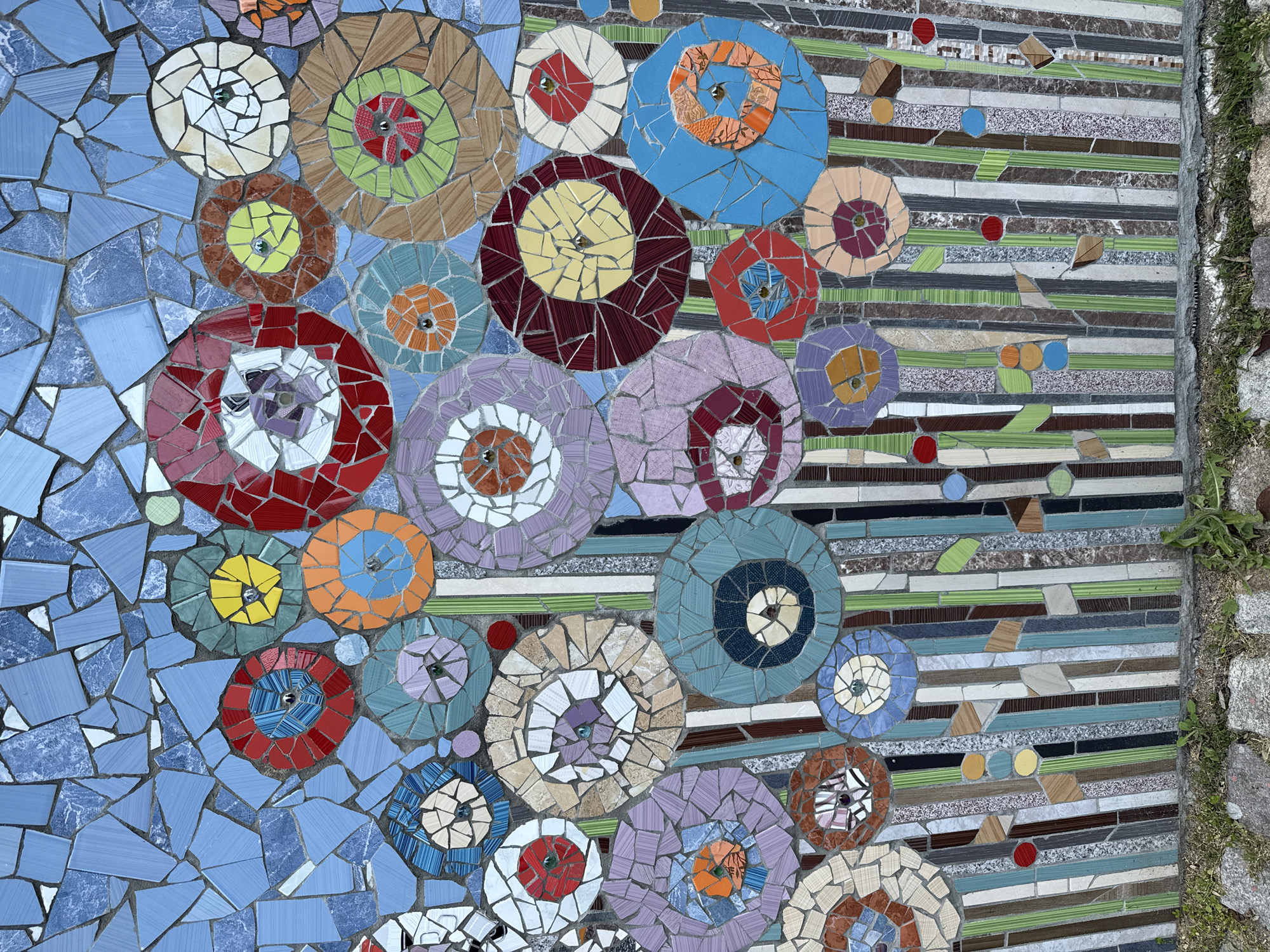 The image shows a section of a mosaic wall in Stara Zagora, Bulgaria. The mosaic consists of colorful, circular flower-like patterns with centers of various colors and materials, surrounded by tiles of different hues and textures. The flowers are set against a backdrop of smaller, irregularly shaped blue tiles on one side and a pattern of horizontal stripes composed of assorted tiles on the other side. A concrete edge with some vegetation growth borders the right side of the mosaic. The mosaic is located outdoors. No people are visible.