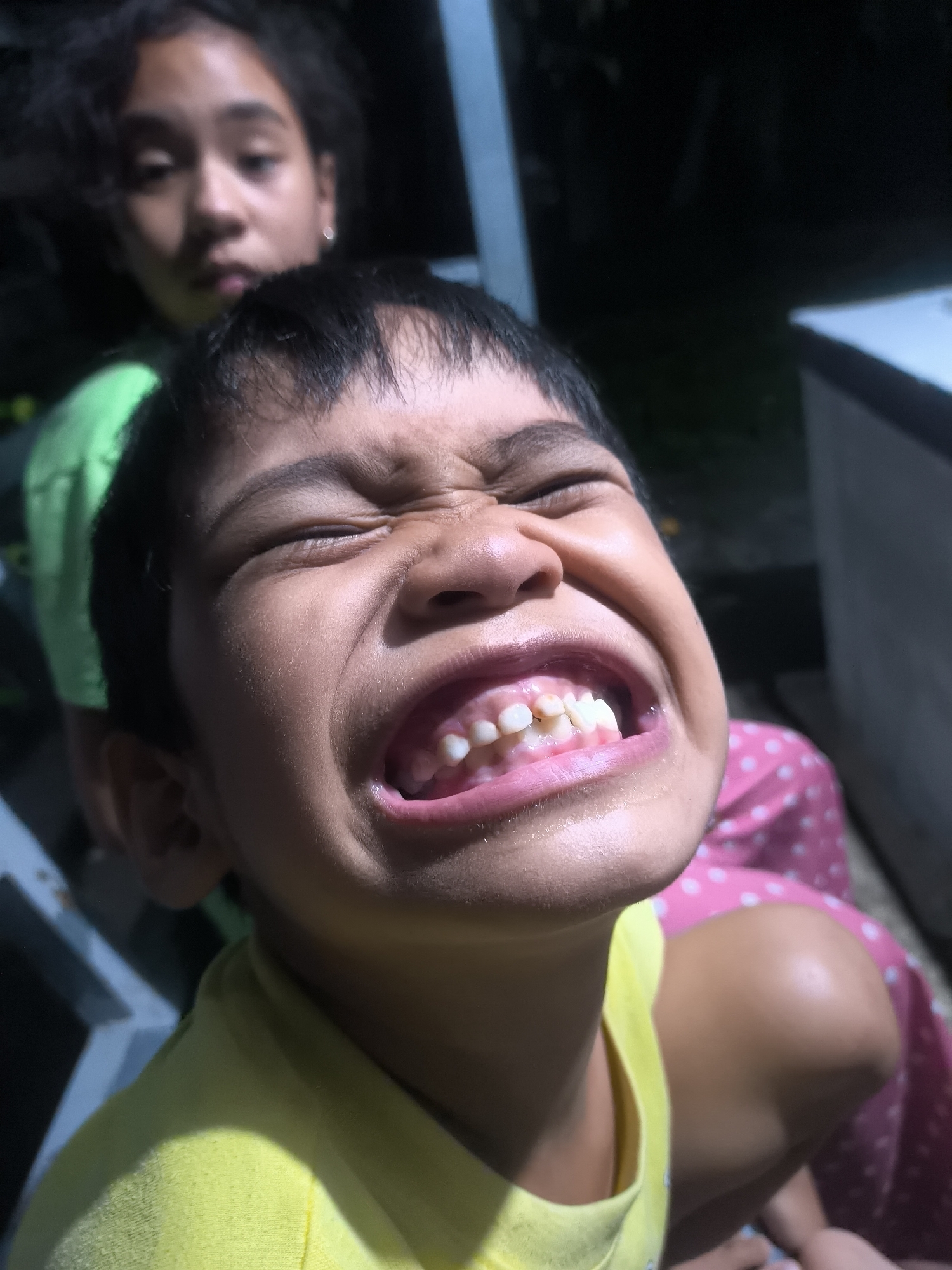 The image features two children, a young boy prominently in the foreground and an older girl blurred in the background. The boy, who appears to be of elementary school age, is captured in a close-up, angled shot. He is wearing a bright yellow t-shirt. His face displays a strong, exaggerated expression: his eyes are squinted shut, his nose is crinkled, and his mouth is wide open, revealing his upper teeth and gums in what could be a forceful laugh, a grimace, or an intense reaction. His dark hair is cut short.
Behind and slightly above the boy, an older girl with dark, wavy hair is visible, out of focus. She is wearing a green shirt, and parts of a pink, polka-dotted garment can be seen near her leg on the right side, suggesting she might be sitting. Her expression is indistinct due to the blur but appears more neutral than the boy's.
The setting is dark and indistinct, creating a high-contrast scene. Strong, directional lighting, possibly natural sunlight, illuminates the boy's face and right shoulder from the side, casting some areas into shadow. In the background, dark shapes and some light-colored structural elements, possibly metal bars or frames, are visible on the left side. A faint white rectangular object can be seen on the far right against the dark backdrop. There is no visible text. The overall scene conveys a candid and energetic moment.