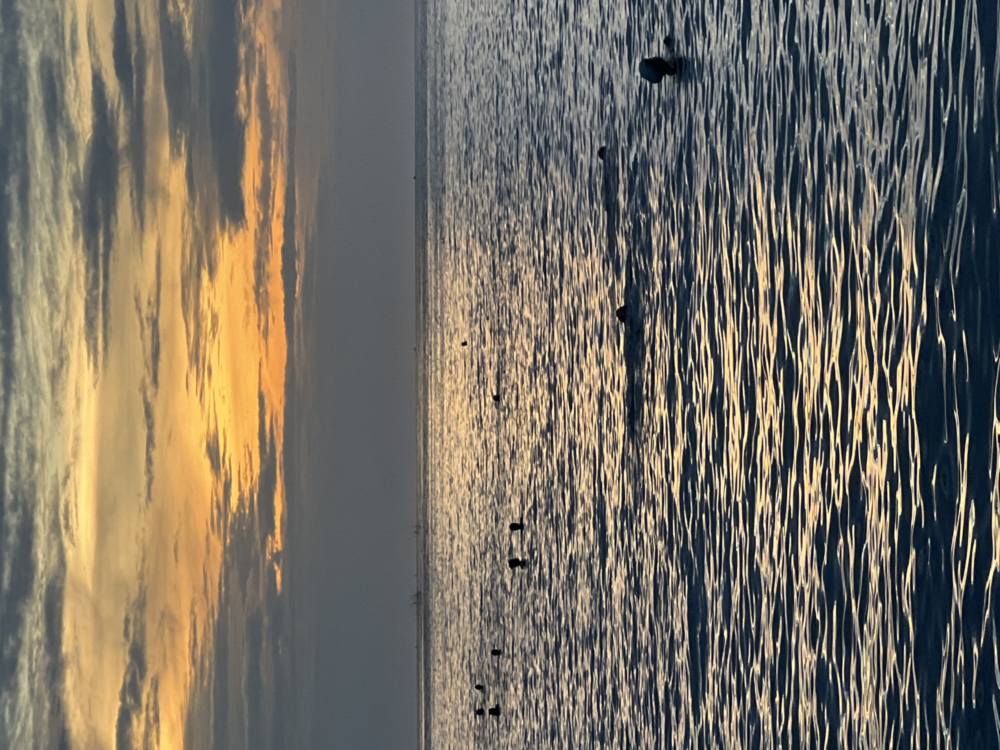 The image depicts a tranquil ocean scene at sunset, with a vibrant sky painted in hues of orange and yellow, contrasting with the darker, cloud-laden upper atmosphere. The water's surface is rippled, reflecting the warm glow of the setting sun. Several figures are visible in the water, appearing to be swimming or wading, with only their heads and shoulders emerging from the surface. The distant horizon shows a faint line of land or other structures, possibly boats or buildings. The overall atmosphere is serene and peaceful, capturing a moment of natural beauty at the close of day. Given the visual cues, this scene could be located along the coast of Nha Trang, Vietnam, a city known for its beautiful beaches and vibrant sunset views.