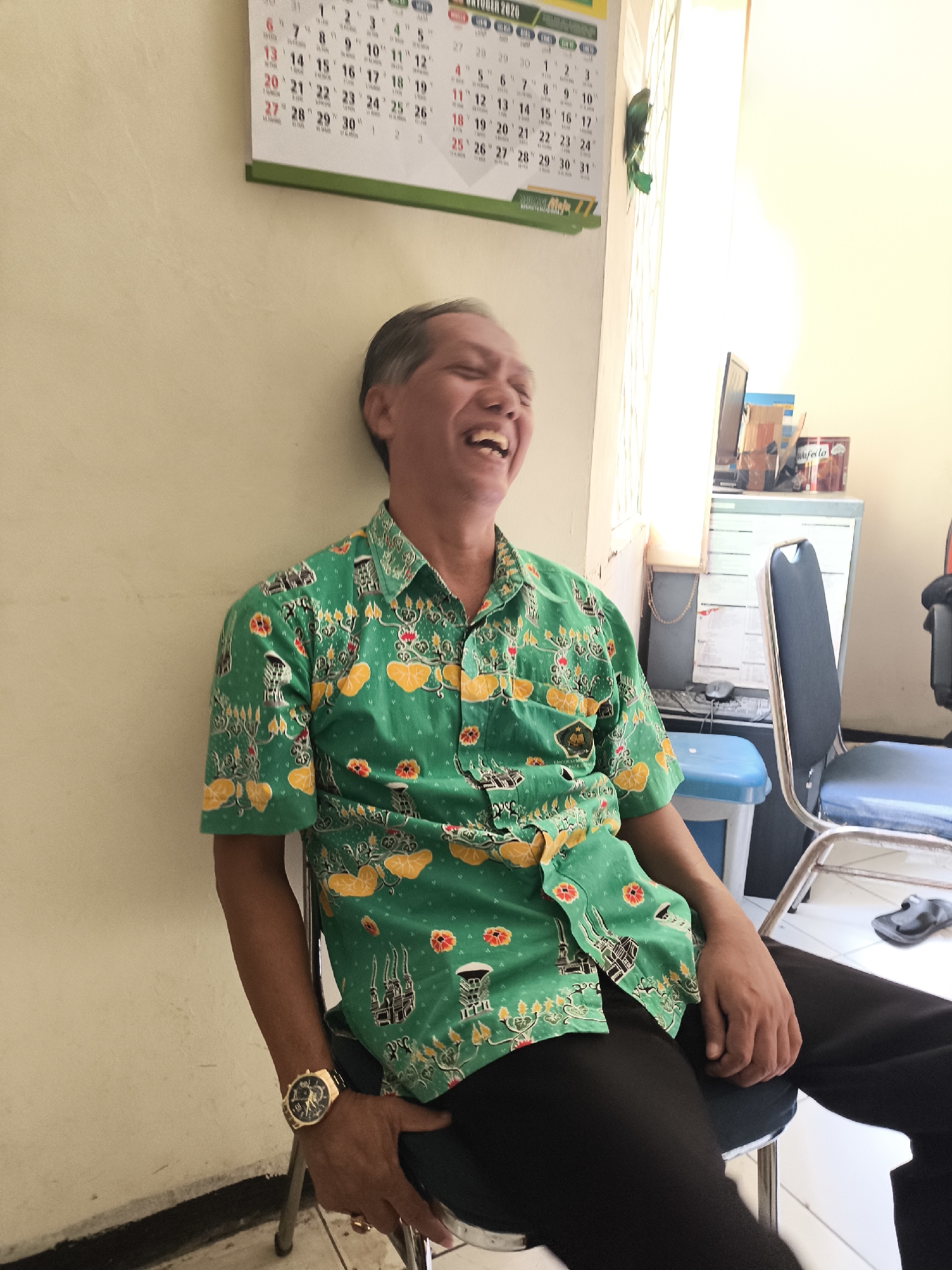 A man with short, dark hair is sitting in a chair against a light-colored wall. He is wearing a bright green short-sleeved button-up shirt with a repeating pattern of yellow and orange floral motifs and black architectural structures. He is laughing with his eyes closed and his mouth open. On his left wrist, he wears a gold-colored watch with a black strap and a dark face with Roman numerals. He is wearing black pants. To the left of the man, a calendar hangs on the wall, showing the month of October 2020. To the right of the man, a filing cabinet and a chair are visible, as well as a window with bright light streaming through. The flooring appears to be tiled. The overall setting suggests an indoor office or waiting area.