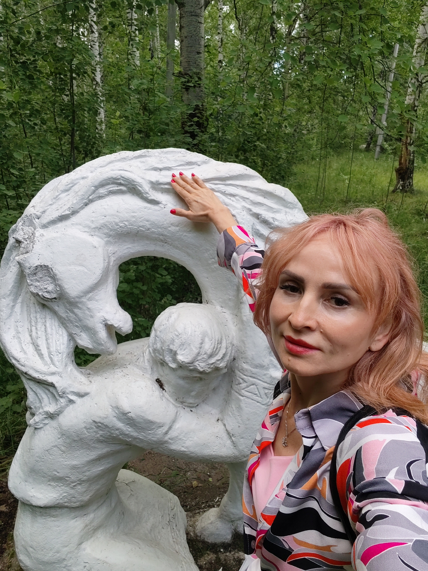 The media file features a woman with peachy-pink hair, wearing a colorful, abstract-patterned shirt and a pink dress underneath, with a delicate necklace. She is posing for a selfie in front of a large, white, abstract sculpture. Her right hand, with bright red nail polish, is placed on the sculpture. The sculpture appears to depict a dynamic scene, possibly involving figures or animals in motion, rendered in a textured, white material.
The setting is outdoors, amidst a lush green forest. The background is filled with dense foliage, including trees with white bark, characteristic of birch trees, and other green undergrowth. The lighting suggests it is daytime, with natural light filtering through the leaves, creating a soft illumination. The overall mood is relaxed and engaged, with the woman smiling gently at the camera. There are no visible text elements in the image. The location context provided indicates this scene is in Chaikovsky, Russia.