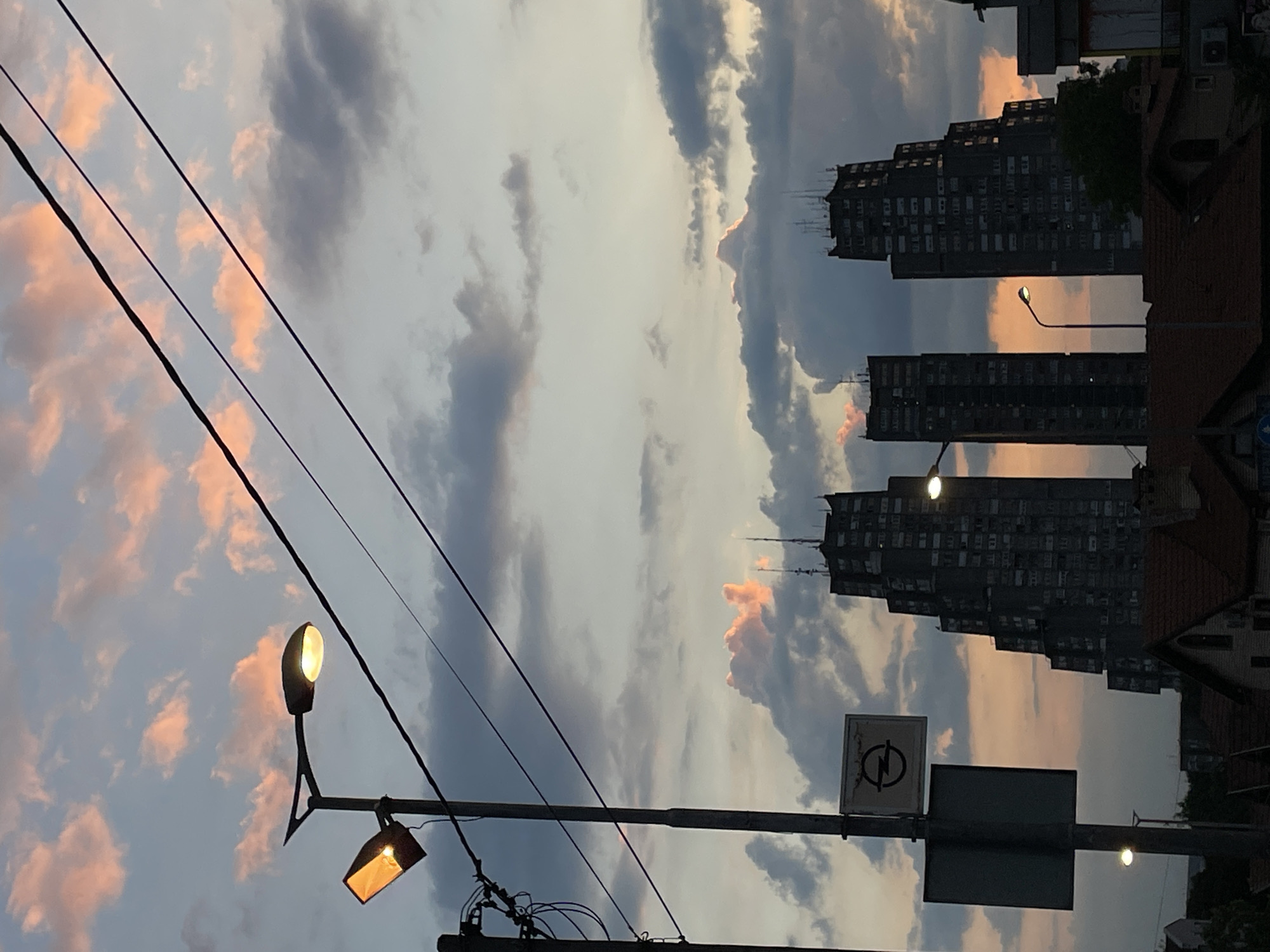 The image depicts a Belgrade city street at dusk. In the foreground, there are utility poles with streetlights, power lines, and a traffic sign. The sky is filled with clouds tinted with pink hues from the setting sun. In the background, three high-rise apartment buildings are visible. They are commonly referred to as "Western City Gate" or "Rudo".