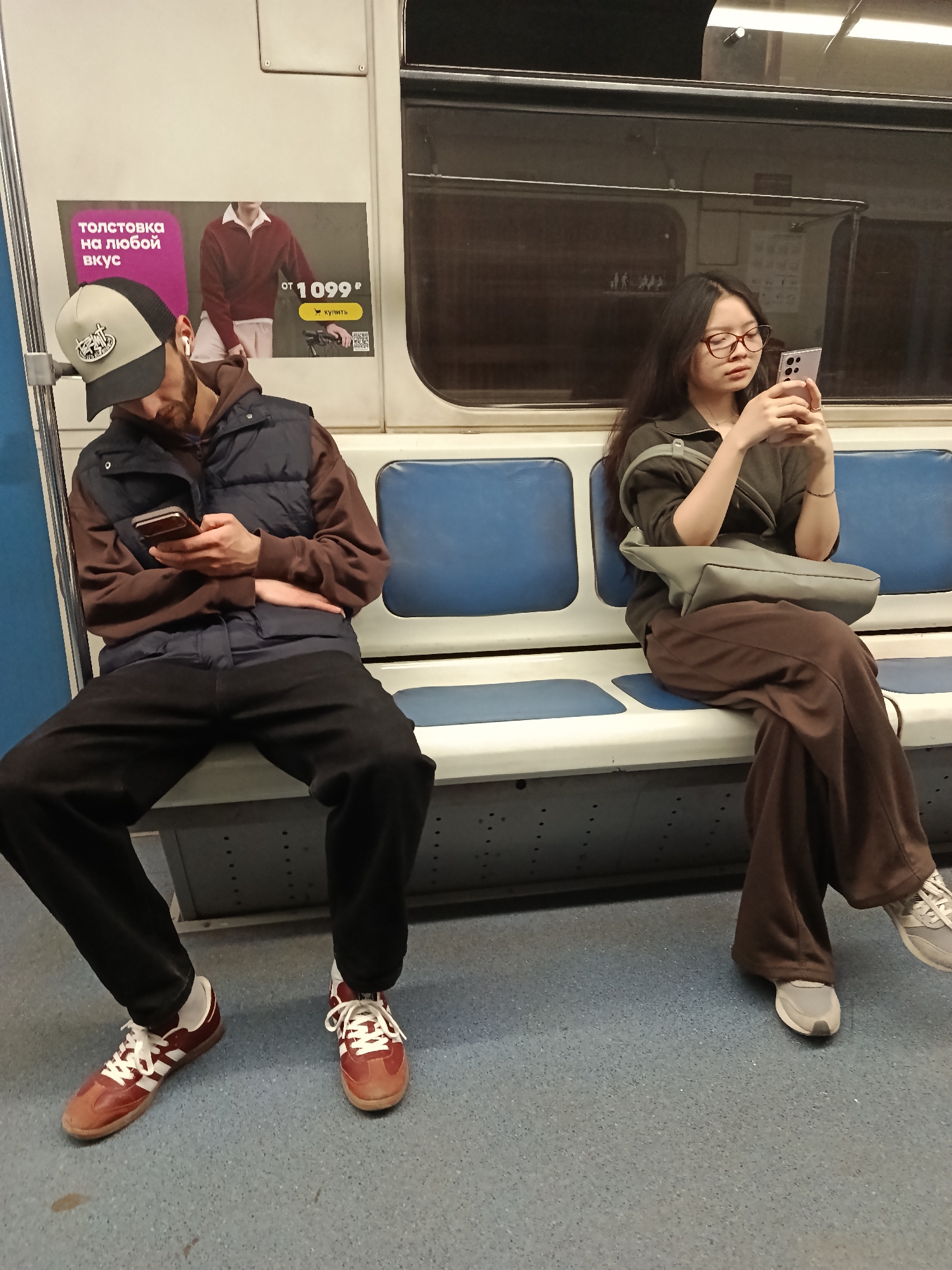 The image captures two individuals sitting on opposite sides of a subway car, both engaged with their smartphones. The setting is a standard transit environment with blue and white seats and a window reflecting the interior of the train.
On the left, a man is seated, wearing a light grey and black baseball cap, a brown hoodie, and a dark blue puffer vest. He has a beard and is wearing white earbuds. He is looking down intently at a dark-colored smartphone held in both hands, resting his forearms on his knees. His attire includes black pants and reddish-brown sneakers with white stripes and laces.
Across from him, on the right, a woman is seated, wearing glasses and a dark green long-sleeved top paired with wide-leg brown pants. Her long dark hair falls over her shoulders. She is holding a light-colored smartphone, possibly pink or lavender, with her hands clasped in front of her, and is actively looking at the screen. A light grey shoulder bag rests on her lap and over her arm. She is wearing light grey sneakers.
An advertisement poster is visible on the wall behind the man. The text is in Cyrillic, reading "ТОЛСТОВКА на любой вкус" (HOODIE for every taste) and displaying a price of "от 1099₽" (from 1099 Russian Rubles). Below the price, there's text "КУПИТЬ" (BUY) and a QR code. The presence of Cyrillic text and the Ruble symbol suggests the location is likely in a Russian-speaking country. Both individuals appear focused on their personal devices, illustrating a common scene of modern commuters.