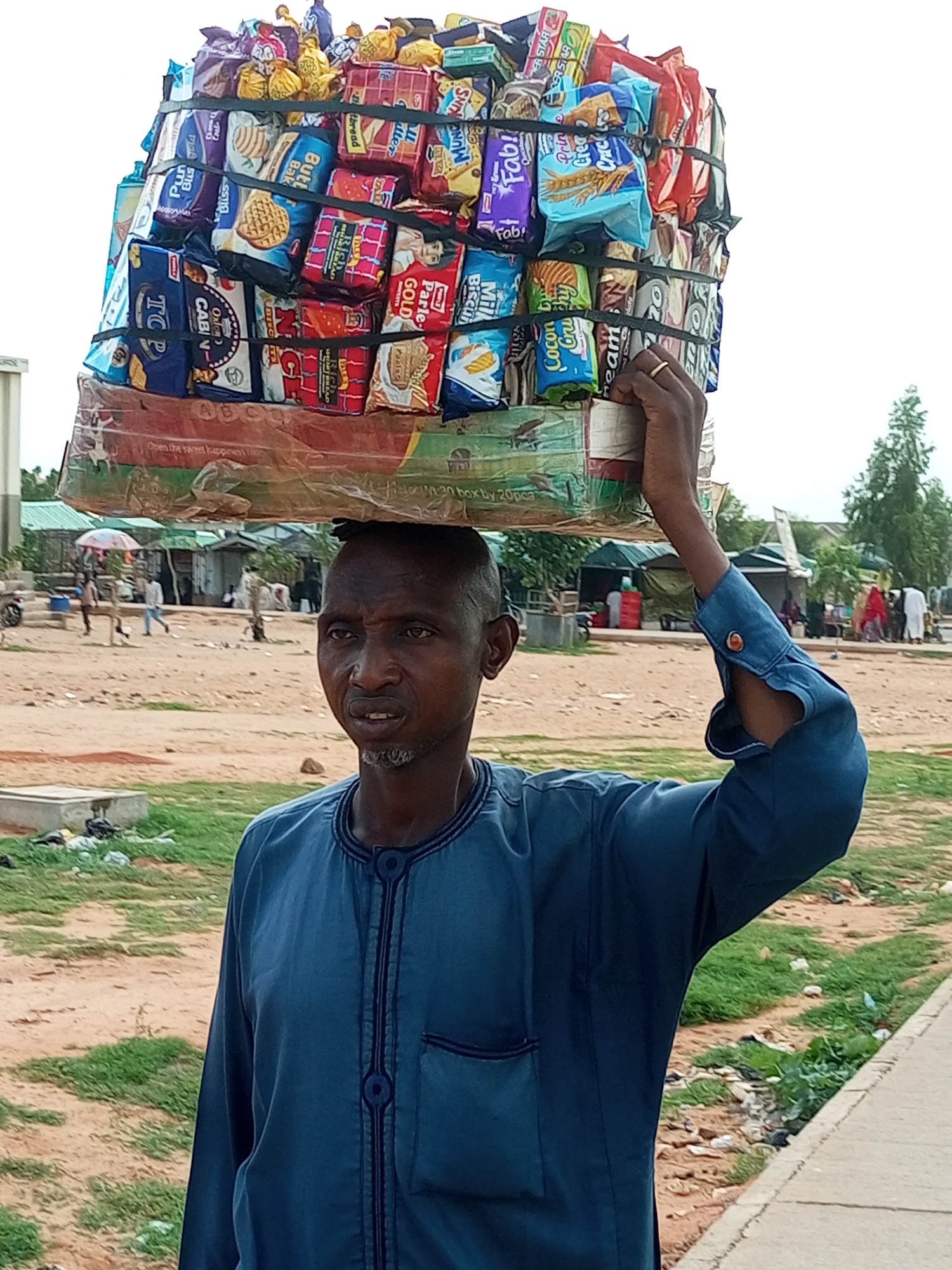 This media file shows a man in Gombe, Nigeria, carrying a large stack of packaged snacks on his head. The man is wearing a blue, long-sleeved garment with intricate dark blue embroidery around the collar and down the front. He has a light beard and appears to be in his middle age.
The stack of snacks is tightly bound with black straps and consists of various brands and types of biscuits and crackers, including packages labeled "Fab!", "MUNCH", "Butt", "Bak", "Bliss", "Parle-GOLD", and "ROCKETS". Some of the packaging is in English, while others are in local languages. The phrase "Open the sweet happiness lice" and "W-30 box by 20pca" are visible on the lower layers of the package.
The setting appears to be an outdoor market or busy street. The ground is a mix of dirt and patchy grass. In the background, there are several small stalls or shops with awnings and people milling about. A few trees are visible, and the sky is overcast, suggesting it might be a cloudy day or early morning/late afternoon. The man's expression is neutral, focusing on the task of balancing the heavy load. The overall impression is of a daily commerce scene in a Nigerian town.