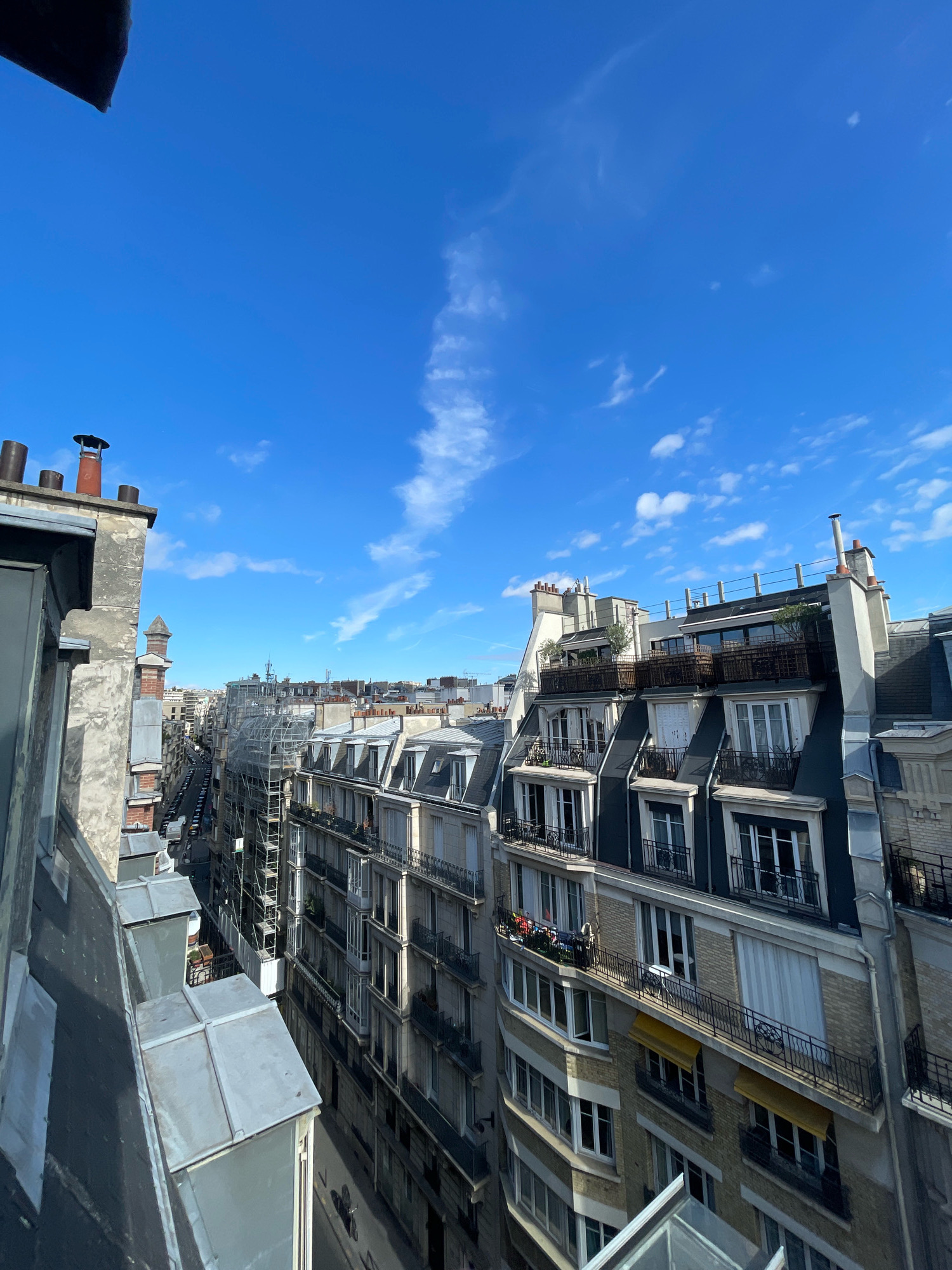This is an eye-level, medium shot of a city street in Paris, France, taken on a clear day. The view is from a building's rooftop, looking down a street lined with typical Parisian Haussmannian apartment buildings. The buildings feature mansard roofs, dormer windows, and decorative iron balconies. Many of the buildings have chimneys. One building on the right has yellow awnings over some windows. On the left, the foreground shows the edge of another building's rooftop with a slanted metallic roof and some pipes. In the middle ground, a building undergoing renovation is covered in scaffolding. The sky is a vibrant blue with scattered white clouds, including a wispy cloud formation stretching across the upper portion of the image. The street below is lined with parked cars, and a few figures can be seen on the sidewalks. The overall impression is a sunny, urban scene.