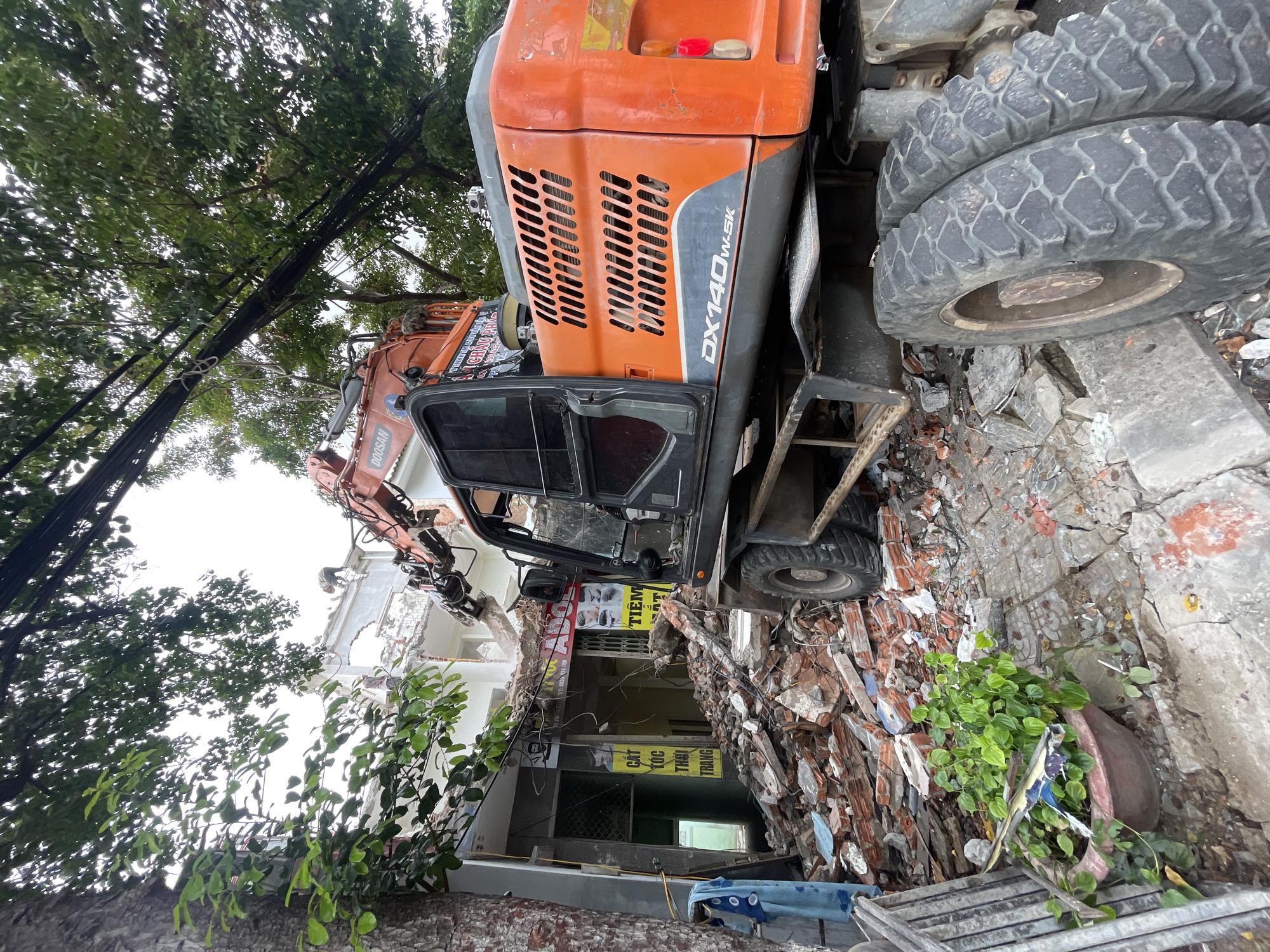 An orange excavator is actively demolishing a building. The excavator's arm is extended, breaking apart the structure. Debris, primarily consisting of bricks and concrete, is scattered across the ground. The building appears to be a storefront with visible signage, including what looks like Vietnamese text and images related to hair salons. A person in work clothes is visible on the roof of the building, near the demolition site. Trees with dense green foliage overhang the scene, and electrical wires are strung across the area. The ground is paved with cracked concrete and rubble. The overall environment suggests an urban demolition site.