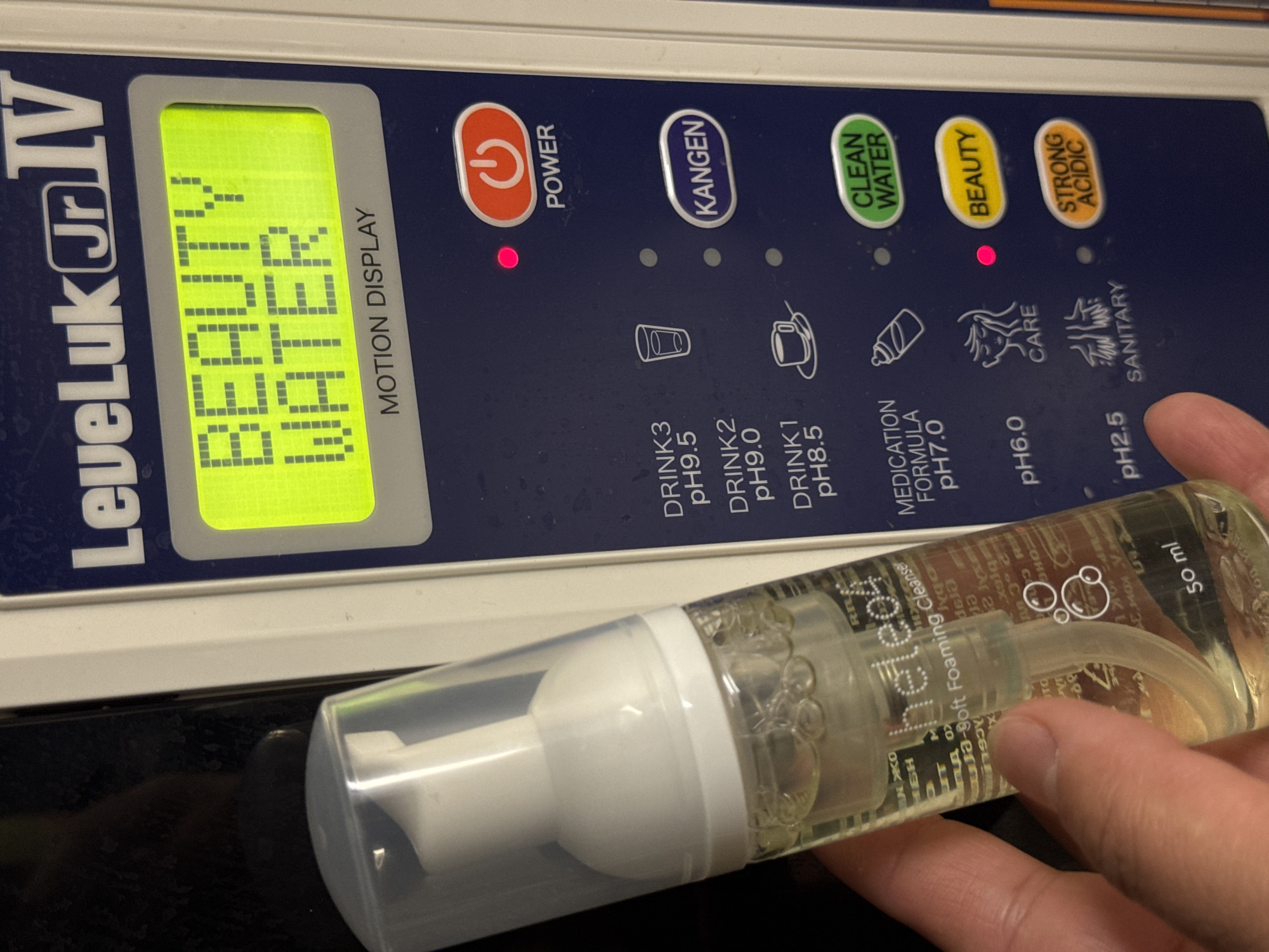 A close-up shot features a "LeveLuk Jr IV" water ionization machine with a digital display showing "BEAUTY WATER." The machine has various button labels including "POWER" (lit red), "KANGEN," "CLEAN WATER," "BEAUTY" (lit red), "STRONG ACIDIC," "DRINK3 pH9.5," "DRINK2 pH9.0," "DRINK1 pH8.5," "MEDICATION FORMULA pH7.0," "pH6.0," and "pH2.5." A clear plastic bottle of "heleo Ja-Soft Foaming Cleanser" is held by a hand in the foreground. The bottle is labeled "50 ml." The environment is generally dark, and the machine appears to be sitting on a black surface. The location is not revealed in this image, so no further location details can be provided.