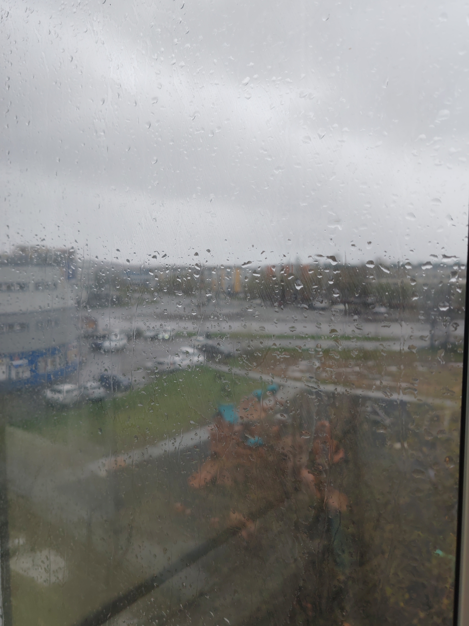The image is a view from a window on a rainy day in what appears to be Petropavlovsk-Kamchatsky, Russia. Raindrops obscure the glass. Beyond the window, a blurred cityscape can be seen, featuring a mix of buildings, roads, parked cars, and greenery. A multi-story building is visible on the left side of the frame. A children's playground and autumn leaves are in the right foreground. The sky is overcast.