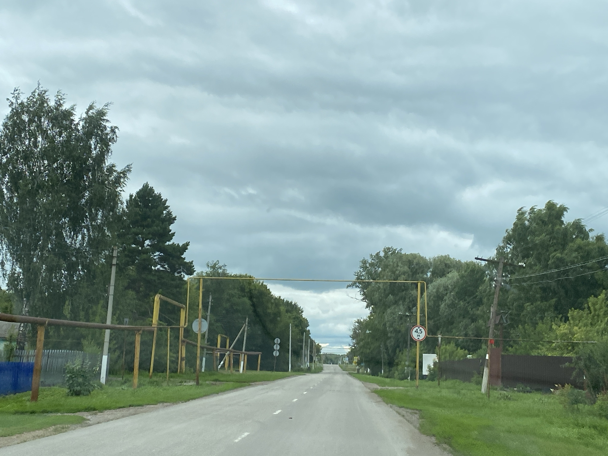 The image depicts a rural road in Nadezhdino, Russia, on an overcast day. The road is paved and has white dashed lines indicating lanes. On either side of the road, there is lush green grass and numerous trees. Along the left side of the road, a blue fence is visible, followed by a brown metal pipeline supported by yellow posts and other rusted metal pipes. On the right side of the road, a brown wooden fence runs parallel to the road, and a power pole with wires is present. Further down the road, a yellow pipe structure spans across the road, with a circular sign indicating a height limit of 4.5 meters. In the distance, the road leads into a more densely wooded area, and the sky is filled with heavy, gray clouds. No people or vehicles are visible in the image.