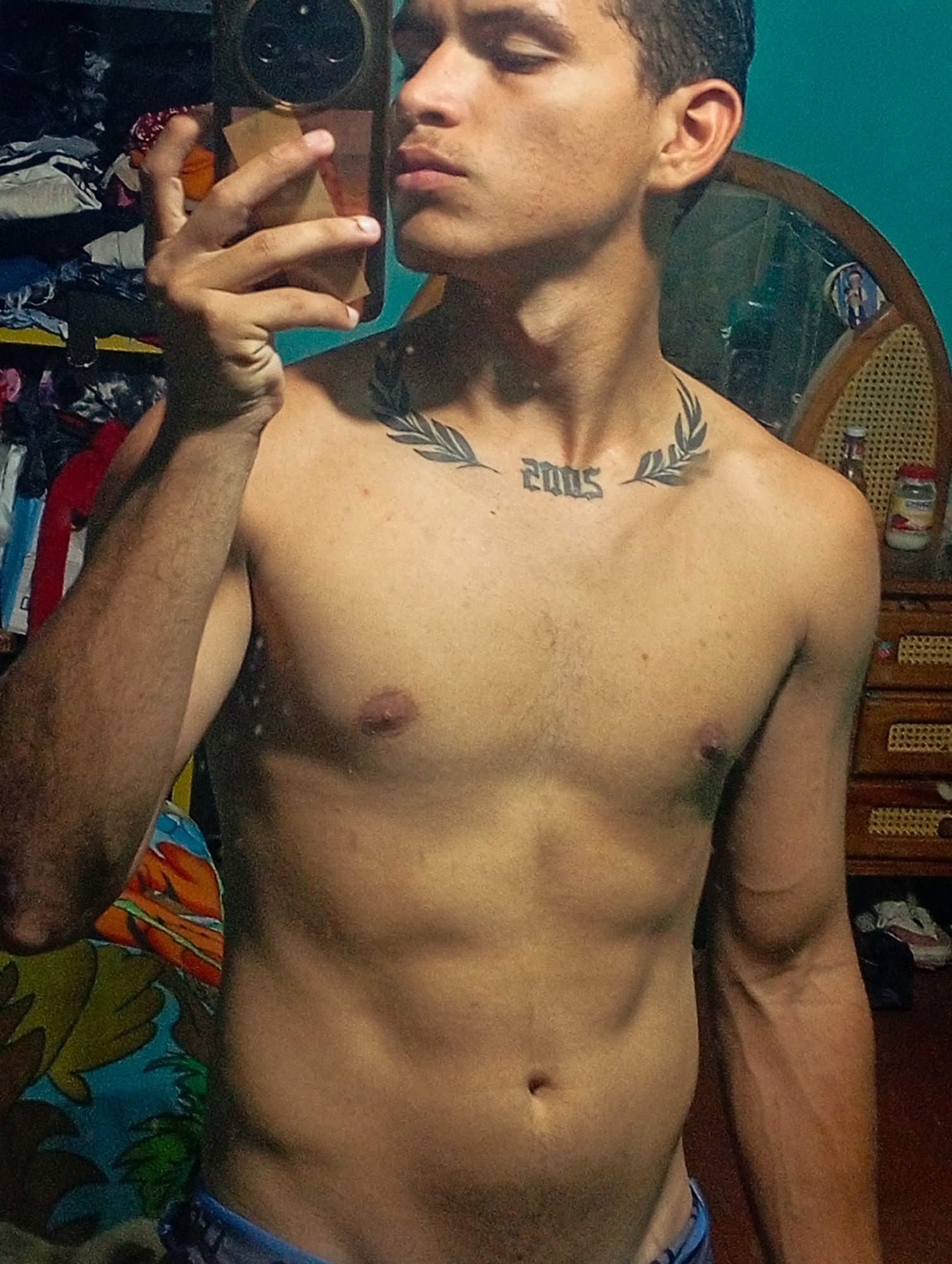 A shirtless young man with a "2005" tattoo and laurel leaves above it on his upper chest is taking a selfie. The background includes a cluttered closet, a mirror, a wooden dresser with items on top, and a teal wall. It's hard to determine if the location is in Altagracia, Venezuela without additional information.
