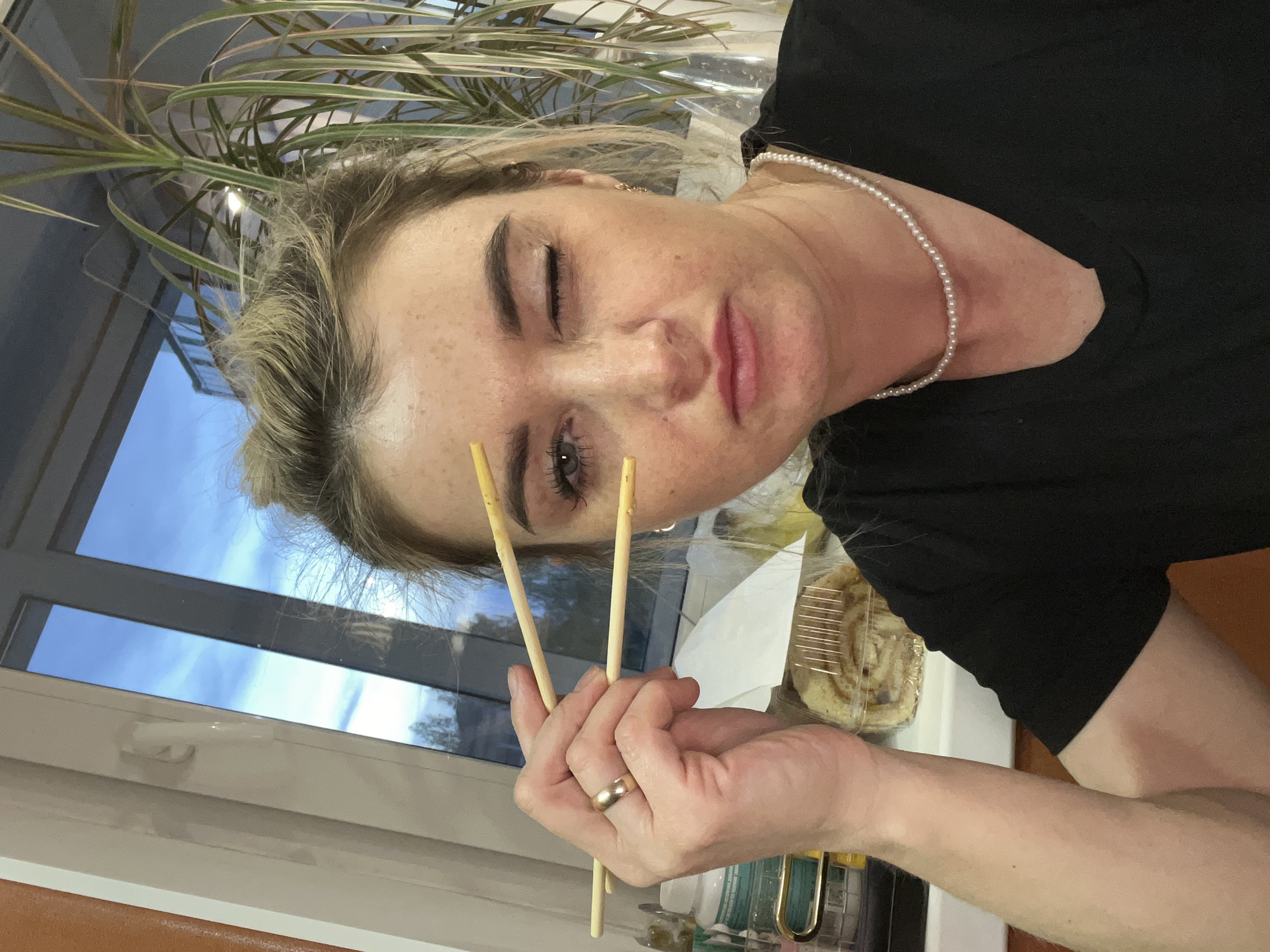 A woman is indoors, holding chopsticks up to her face, possibly in Chaikovsky, Russia. She has light skin, blonde hair pulled back, and is wearing a black shirt and a pearl necklace. Her left eye is closed in a wink. She is wearing a gold ring on her left ring finger. In the background is a window showing a sky with clouds, a plant in a pot, and part of a pastry in a clear container. Part of a kitchen counter is visible in the foreground.
