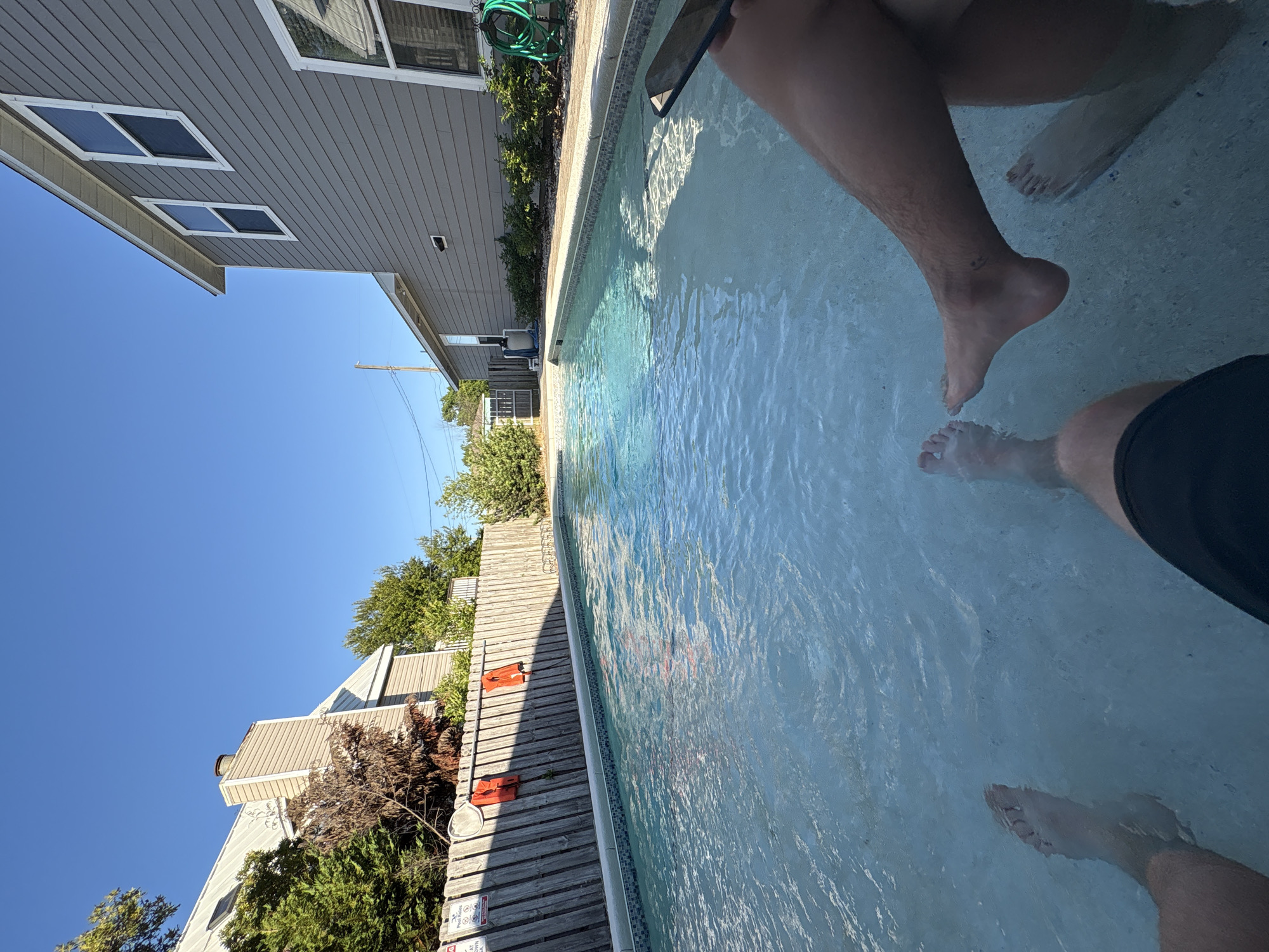 The image captures a partial view of a swimming pool, with multiple people's legs and feet immersed in the water. The pool is bordered by a wooden deck and overlooks residential buildings. The sky is clear and blue. The scene likely depicts a casual, relaxed setting, possibly a backyard or private residence in Panama City Beach.