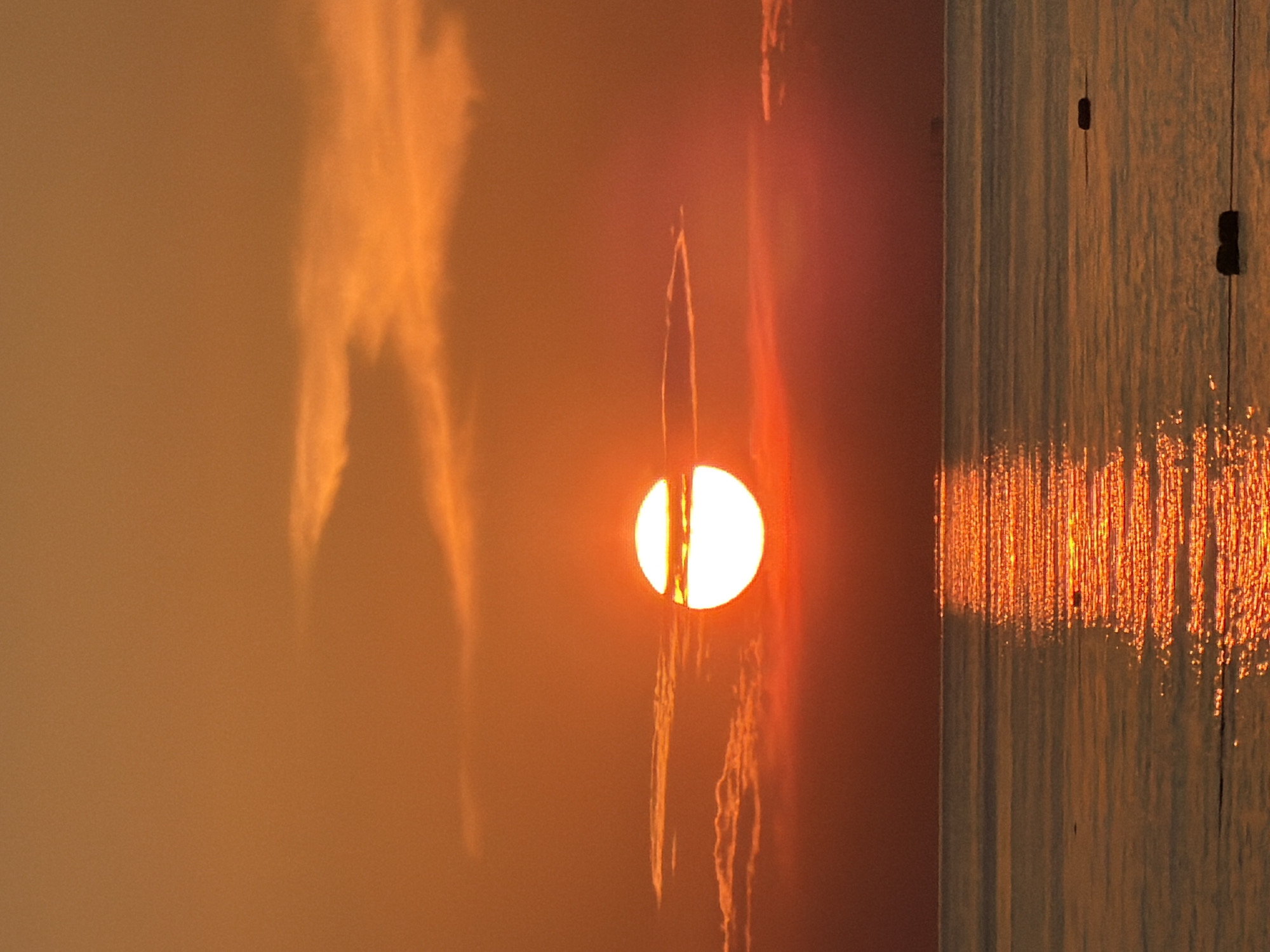 A vertical, medium shot captures a sunset over the Black Sea, possibly near Sochi, Russia. The sun, a bright white orb with a distinct horizontal dark band, is positioned slightly left of center in the middle of the frame. Wispy clouds, illuminated by the sun's orange glow, streak across the sky. The sky transitions from a warm orange at the top to a deep red near the horizon. The calm sea reflects the vibrant colors of the sunset, creating a shimmering path of light. Two dark, rectangular buoys are visible in the water, one in the lower right quadrant and another further to the left and slightly higher. The scene is serene, with no people or discernible human activity other than the buoys.