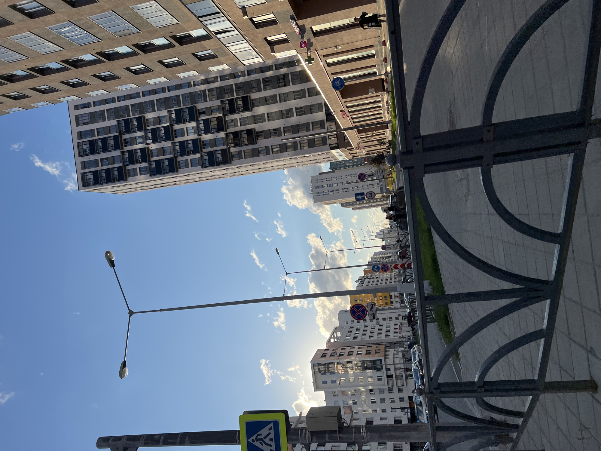 The image depicts a modern urban street scene in Yekaterinburg, Russia, characterized by a row of tall apartment buildings. On the right side of the frame, a dark brown, multi-story building with numerous windows and balconies dominates the view. To its left, a lighter-colored, patterned apartment building of similar height is visible. The street itself is paved and lined with sidewalks. Several streetlights with multiple lamps are positioned along the road. Traffic signs, including a pedestrian crossing sign and speed limit signs, are present. A dark metal fence runs along the foreground of the image, separating the sidewalk from the street. The sky above is blue with scattered white clouds, and the sun is casting a warm light. A few cars are seen on the road, and a person is walking on the sidewalk in the distance. The overall impression is that of a newly developed residential area within the city.