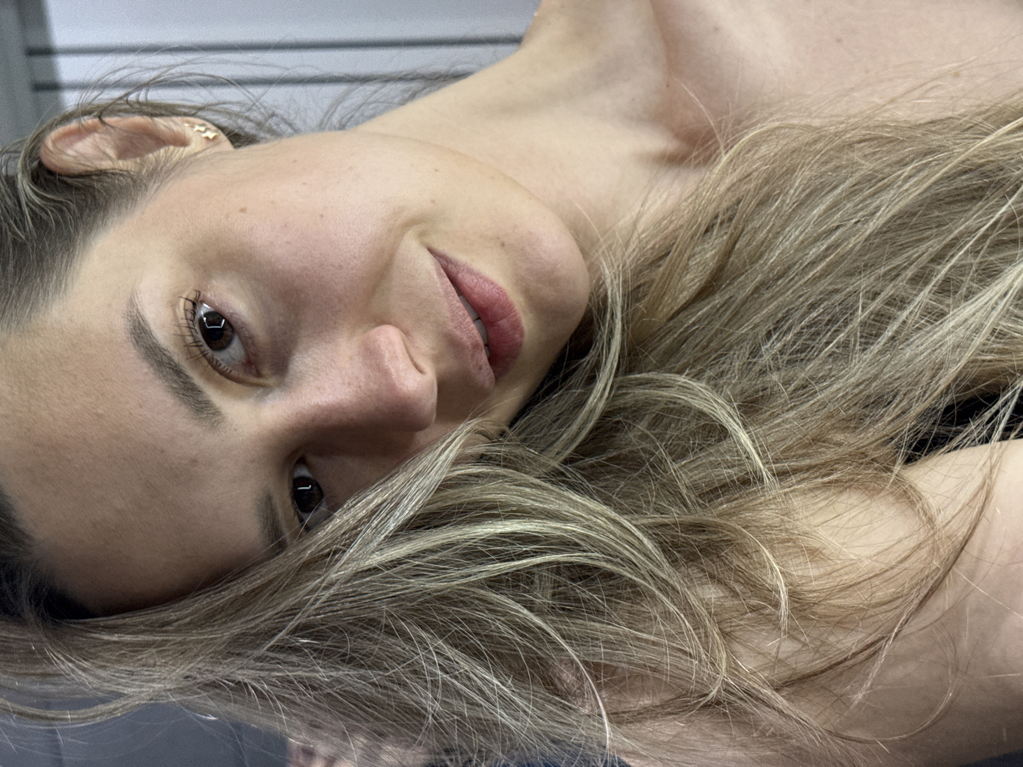 A close-up portrait captures a woman with light brown hair and blonde highlights, positioned with her head tilted, looking directly at the camera. She has brown eyes, well-defined eyebrows and eyelashes, and a subtle, relaxed smile on her lips. Her skin appears natural, with visible pores and slight freckling. A small, gold-toned earring is visible in her right ear.
Her long, tousled hair is spread out, framing her face and resting over her bare shoulders and upper chest, which are visible in the foreground. The background is softly blurred, showing horizontal lines on a light-colored surface, suggesting an indoor setting with either blinds or paneled walls. The lighting is soft and even, highlighting her features without harsh shadows. The overall mood is intimate and serene, conveying a sense of natural beauty and calm. There is no visible text in the image. The location is Chaikovsky, Russia, though this cannot be discerned from the visual context.