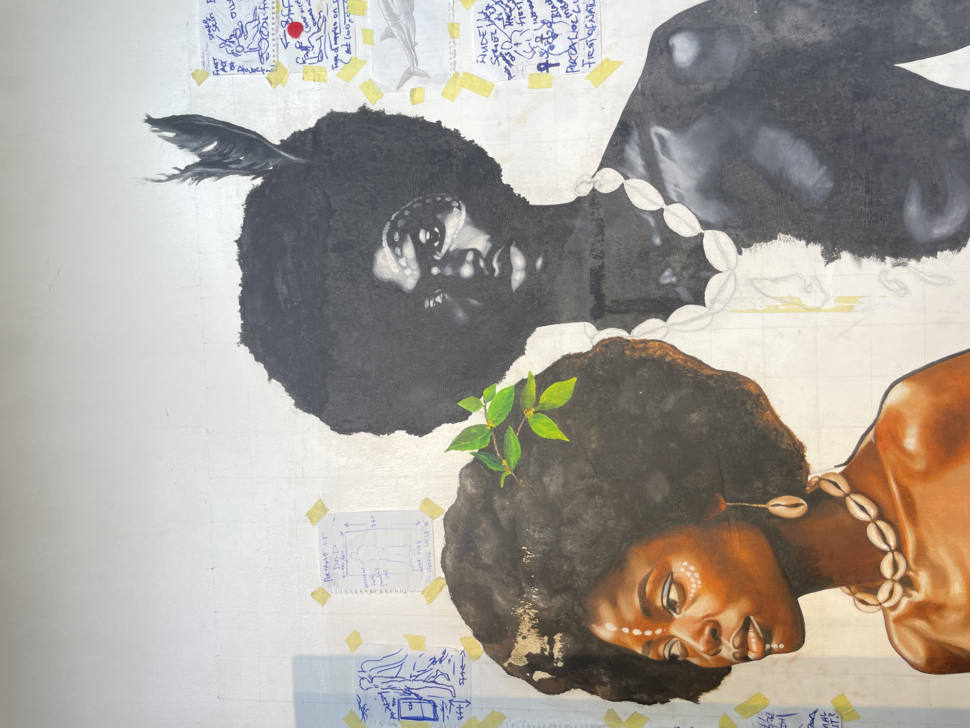 The image shows a work in progress, seemingly an unfinished painting of two African women's portraits on a silver-toned background. The women are painted in different styles: one in grayscale with a feather in her hair and a white bead necklace, and the other in color with a branch of green leaves in her hair and a necklace of cowrie shells. Both have white markings on their faces. The background is covered with taped-up sketches, notes, and diagrams in blue ink. Text fragments visible include "PORTRAIT OF DAP", "State", and references to "FIRST OF NATU". The painting appears to be in a studio or indoor setting. There is no definitive information indicating the location of the city of Ado, Nigeria.