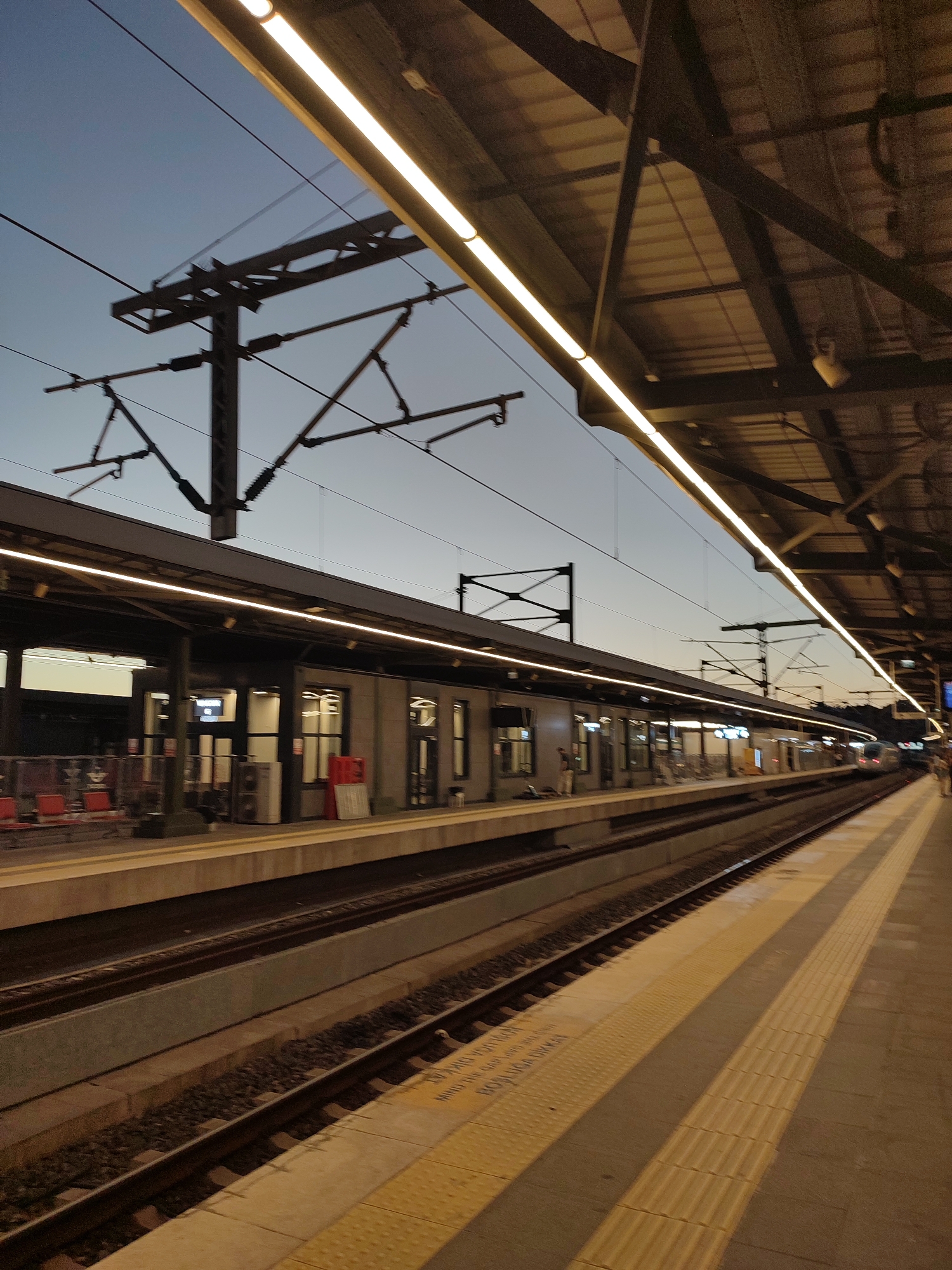 This is a ground-level, eye-level, medium shot of a train station platform at dusk. The sky is a pale blue, transitioning to a darker hue at the top of the frame, with overhead power lines and their support structures visible. The station platform is covered by a large, angular roof supported by dark metal beams. Along the edge of the platform are bright, linear lights. The platform itself features yellow tactile paving near the edge of the tracks. On the left side of the frame, a train car is visible at the platform, with red seating and closed doors. In the distance, on the right side of the frame, another train is approaching the station. The train tracks are laid with ballast. The station appears to be the Söğütlüçeşme station in Kadıköy, Istanbul, Turkey, judging by the architectural style, overhead lines, and the presence of a tram line often associated with the area.